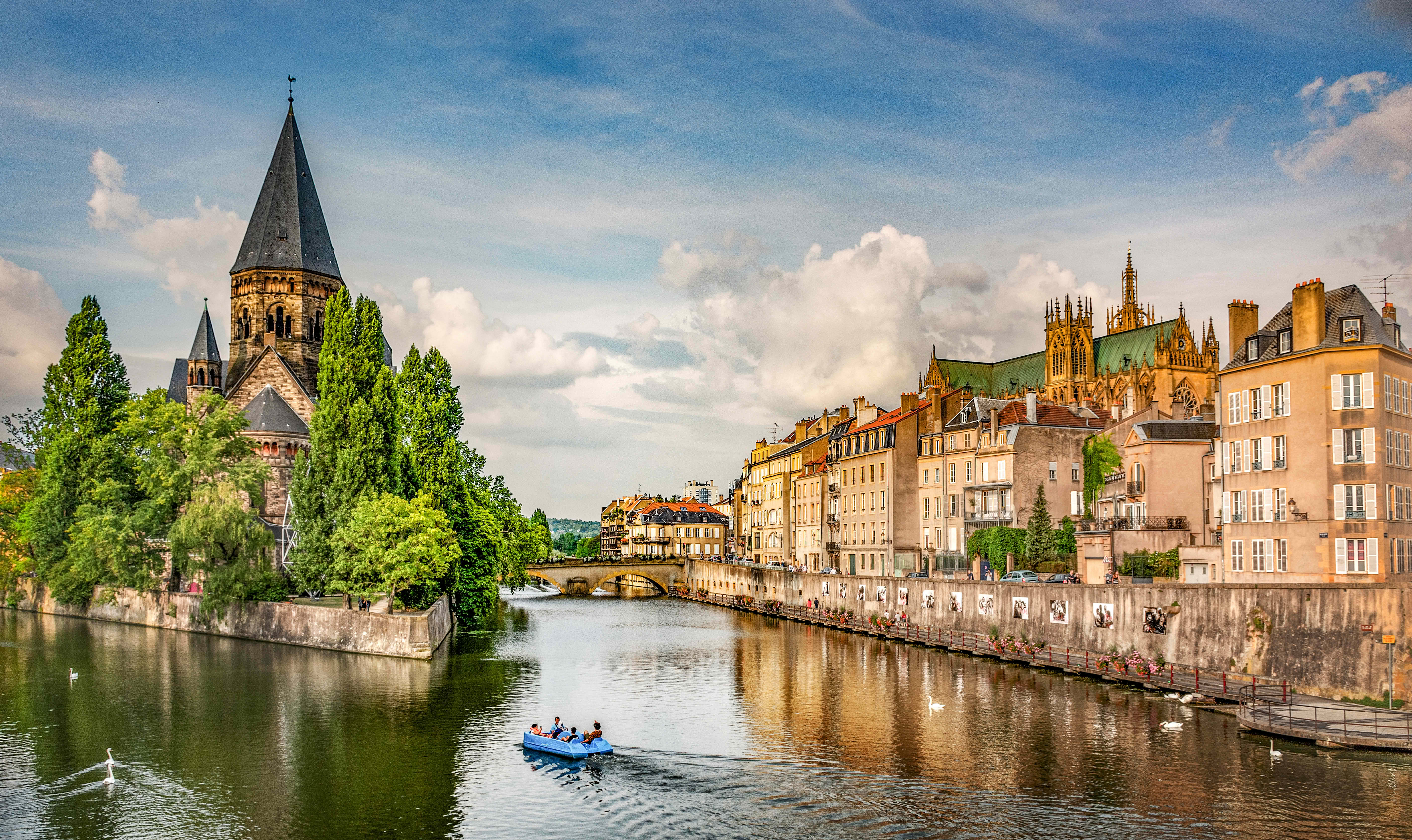 Rivière traversant une ville européenne avec bateaux, cygnes, église à flèche et bâtiments historiques le long des berges.