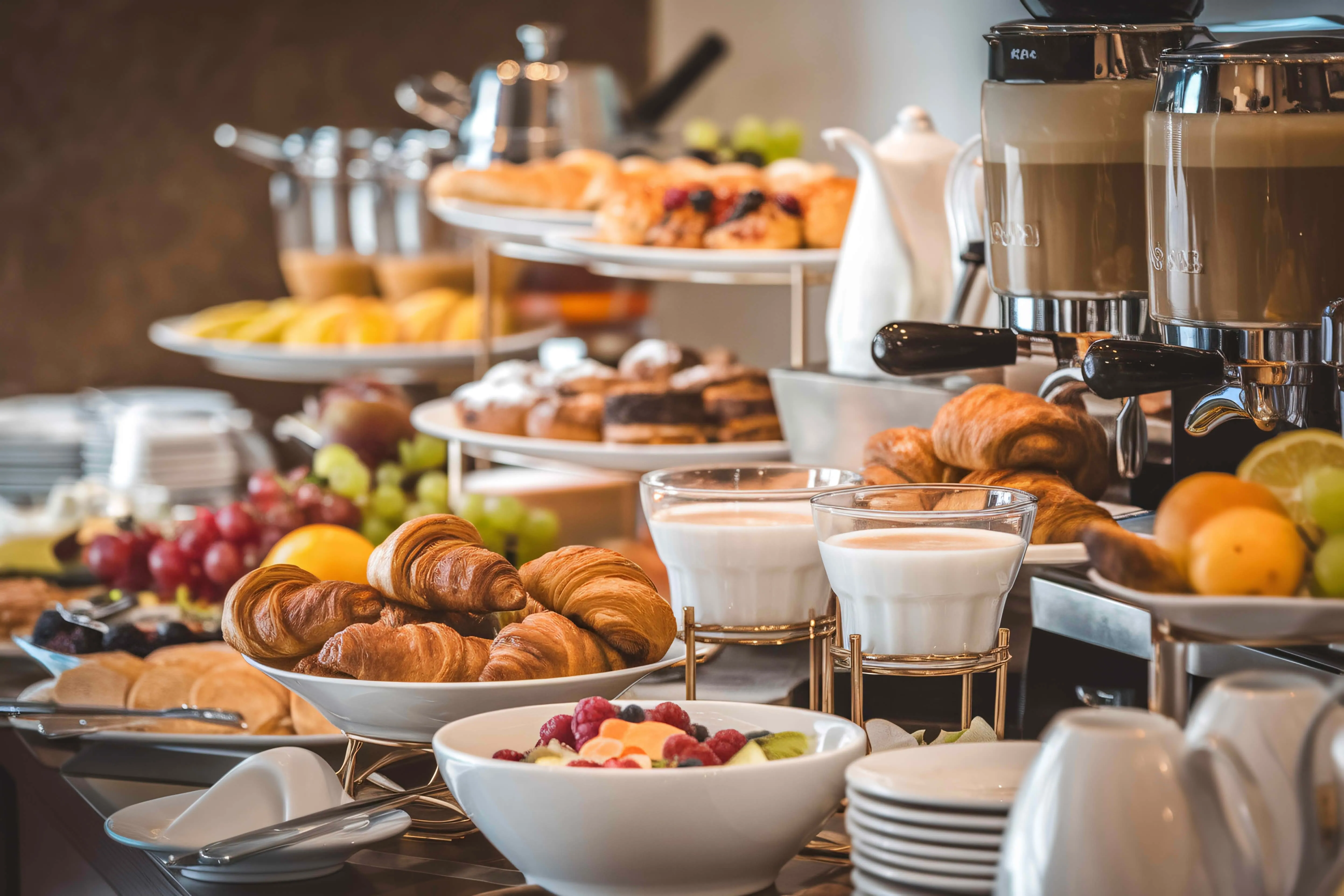 Table avec un assortiment de produits pour le petit-déjeuner, notamment des croissants, des fruits frais, des yaourts, des viennoiseries et une machine à café.