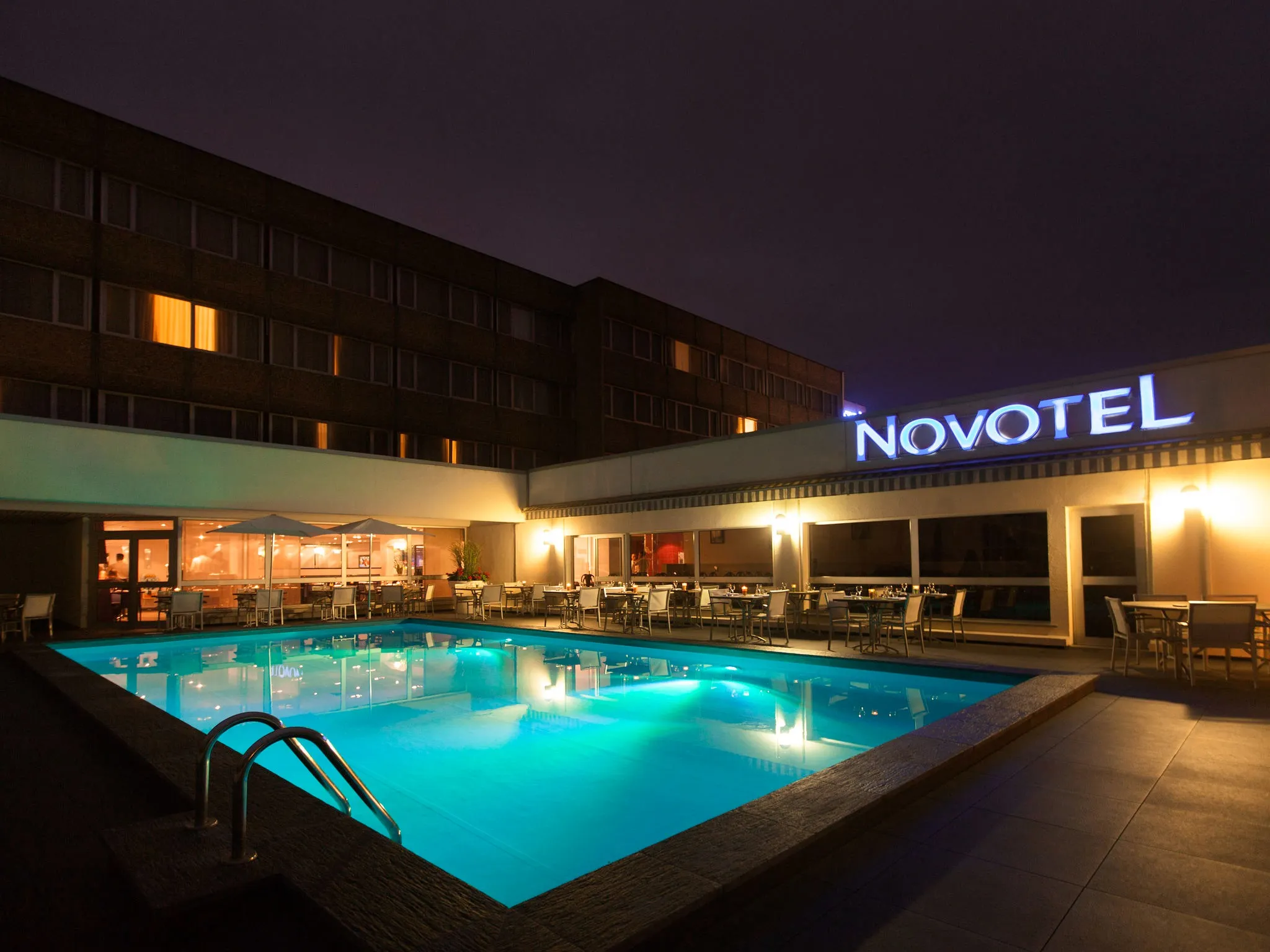Piscine éclairée la nuit avec tables et chaises autour sous un panneau lumineux NOVOTEL.