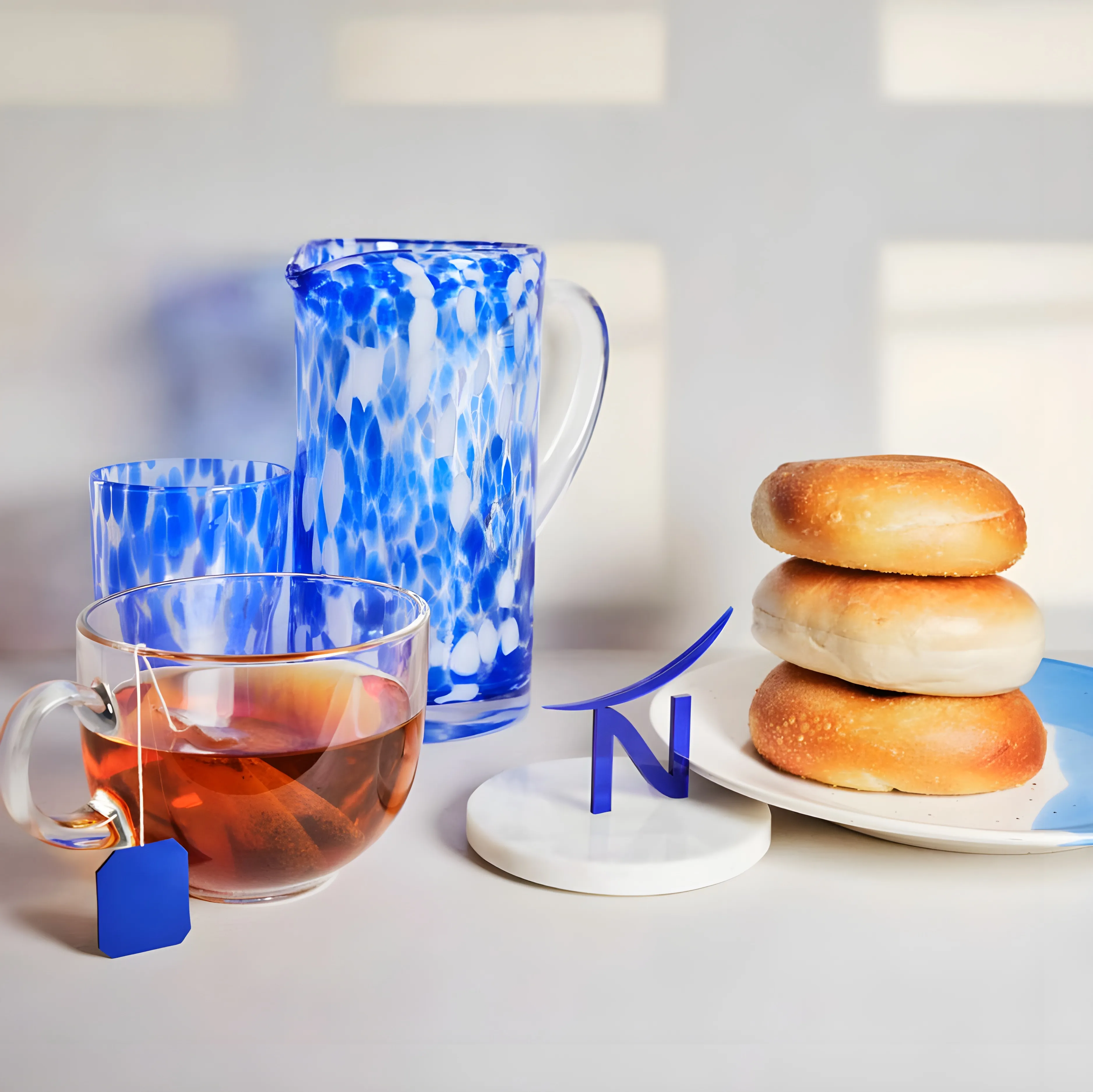 Tasse de thé avec sachet, pichet et verre en verre bleu tacheté, et trois petits pains empilés sur une assiette avec un logo bleu.