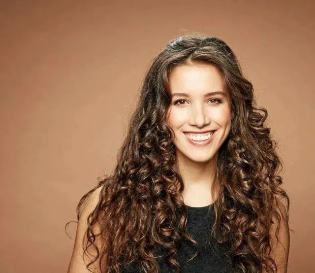 Junge Frau mit langen, lockigen braunen Haaren lächelt vor braunem Hintergrund.