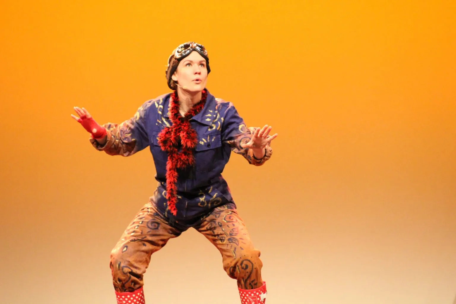 Theaterschauspielerin in bunter Kostümierung mit roten Gummistiefeln und Schal vor orangefarbenem Hintergrund.
