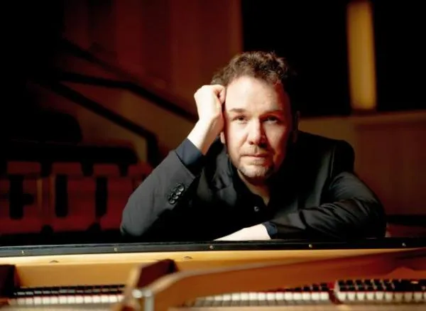 Mann mit dunklem Sakko lehnt an einem Klavier und schaut direkt in die Kamera in einem dunklen Raum.