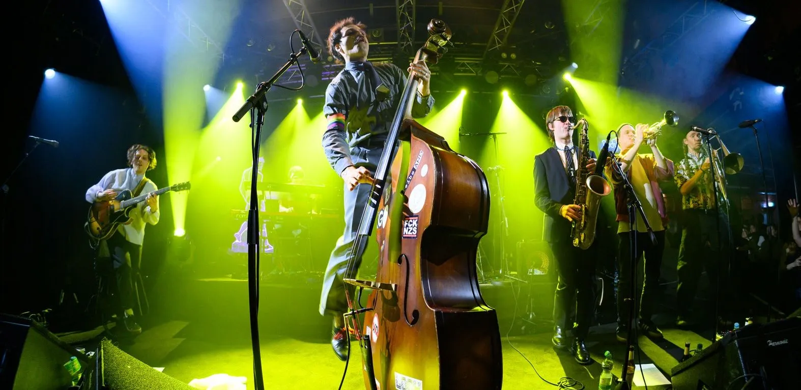 Live band performing on stage with musicians playing double bass, guitar, saxophone, trumpet, and trombone under yellow and blue stage lights.
