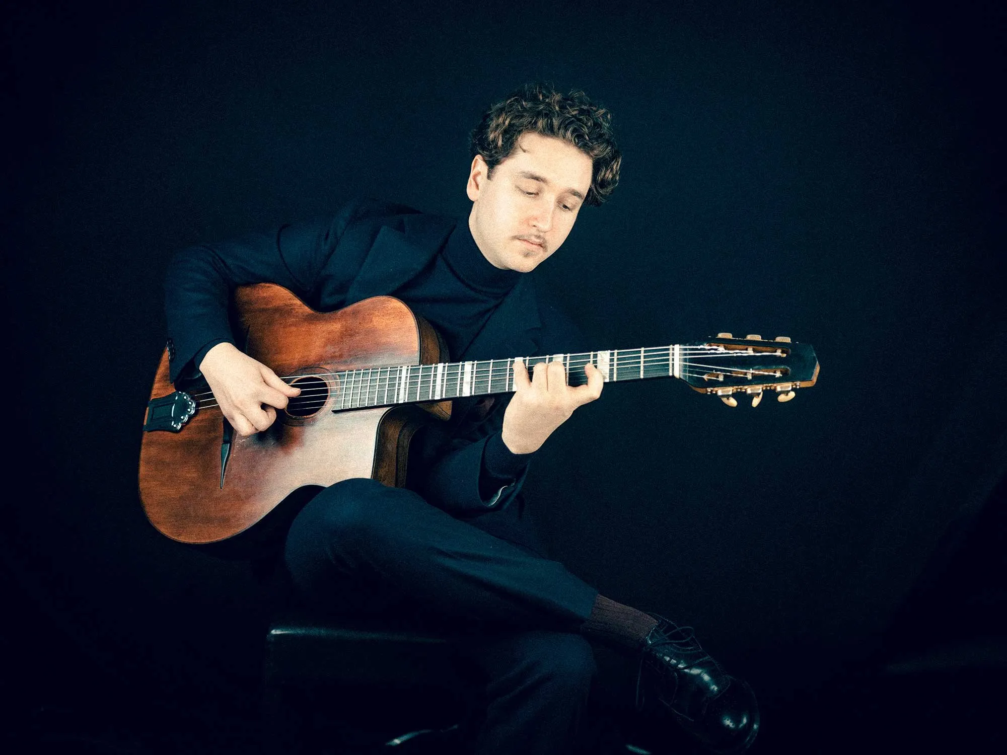 Ein Mann in schwarzer Kleidung spielt konzentriert eine braune akustische Gitarre vor schwarzem Hintergrund.