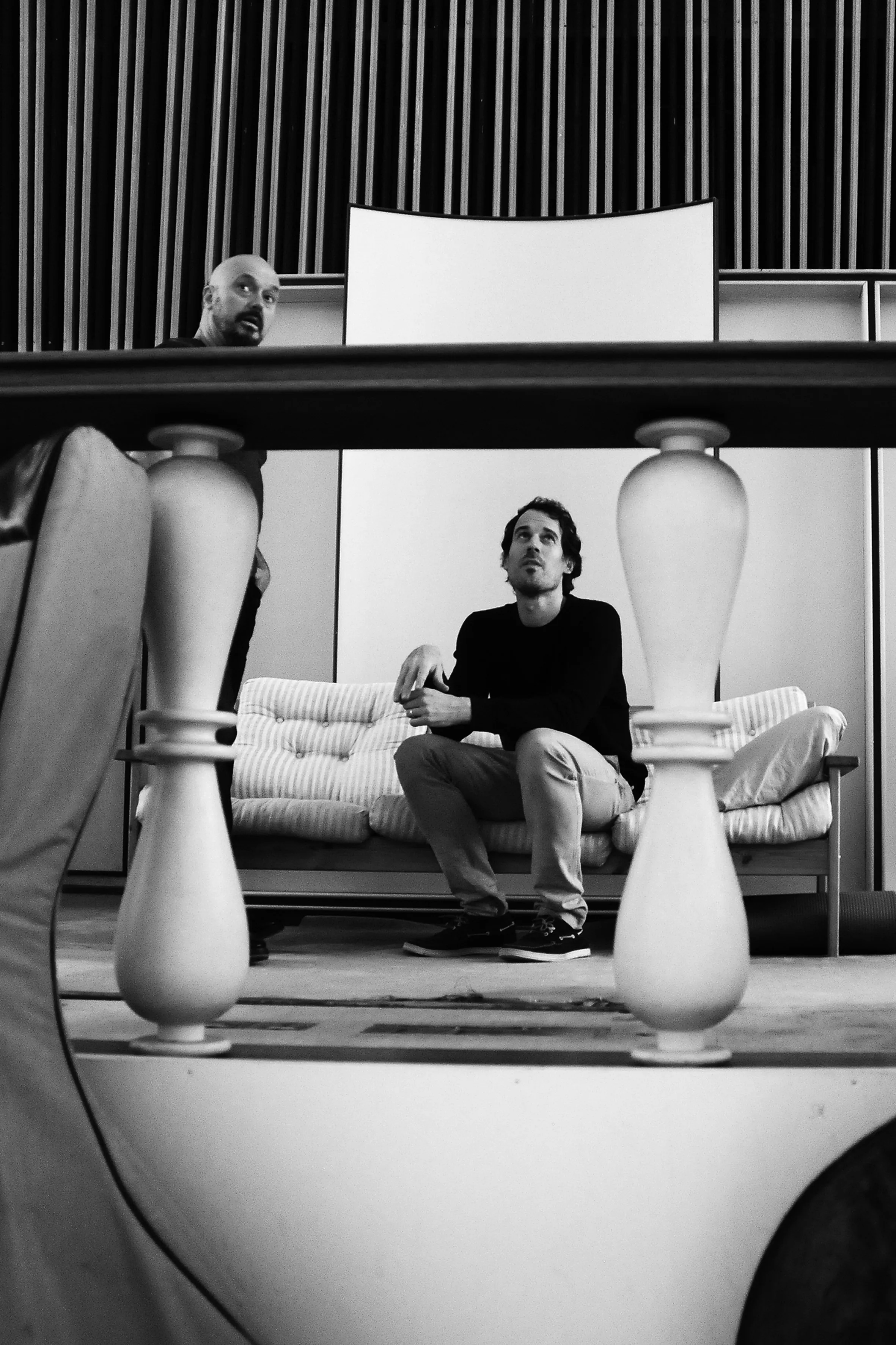 Zwei Männer in einem Raum, einer sitzt auf einem Sofa und schaut nach oben, der andere steht im Hintergrund.