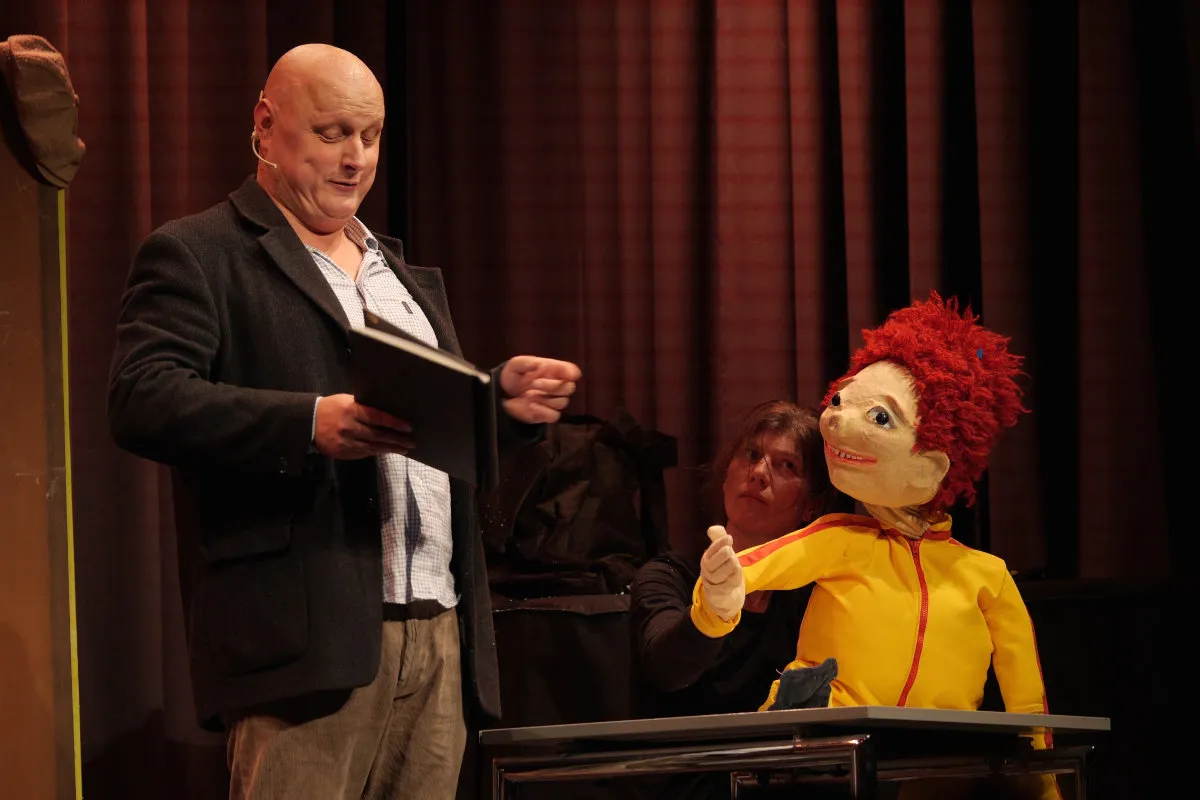 Ein Mann liest von einem Buch, während eine Frau eine rote Handpuppe in gelber Jacke an einem Tisch steuert.
