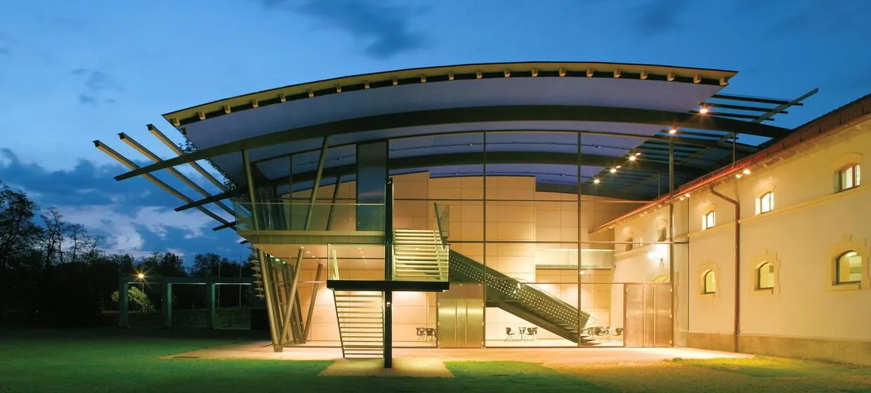 Modernes Gebäude mit großen Glaswänden, beleuchteten Innenräumen und Außentreppen bei Dämmerung.