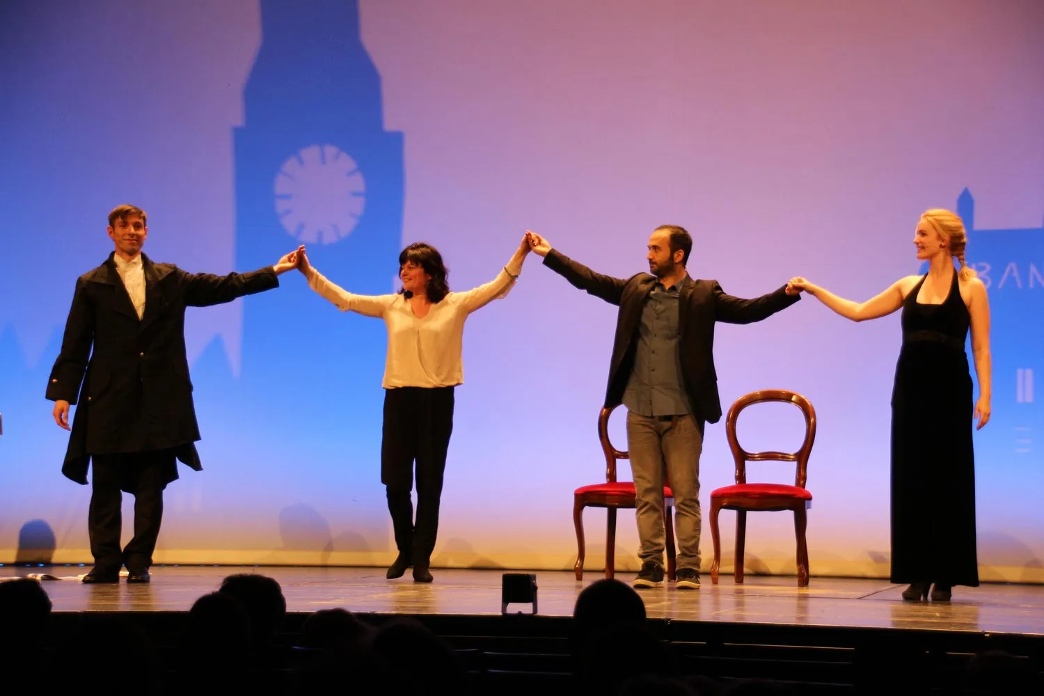 Vier Schauspieler stehen an einer Bühne, halten sich an den Händen, vor einem Hintergrund mit einer großen Schattenprojektion einer Uhr und Gebäuden.