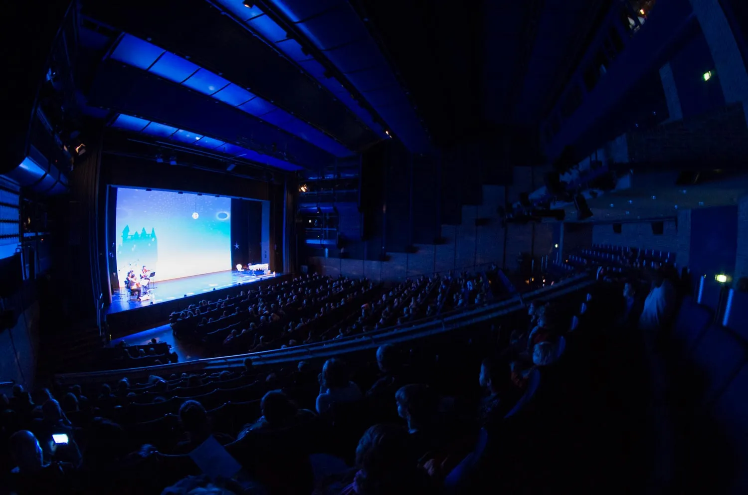 Vollbesetztes Theater mit Bühne, auf der Musiker und eine Projektion mit einem Schloss und Nachthimmel zu sehen sind.