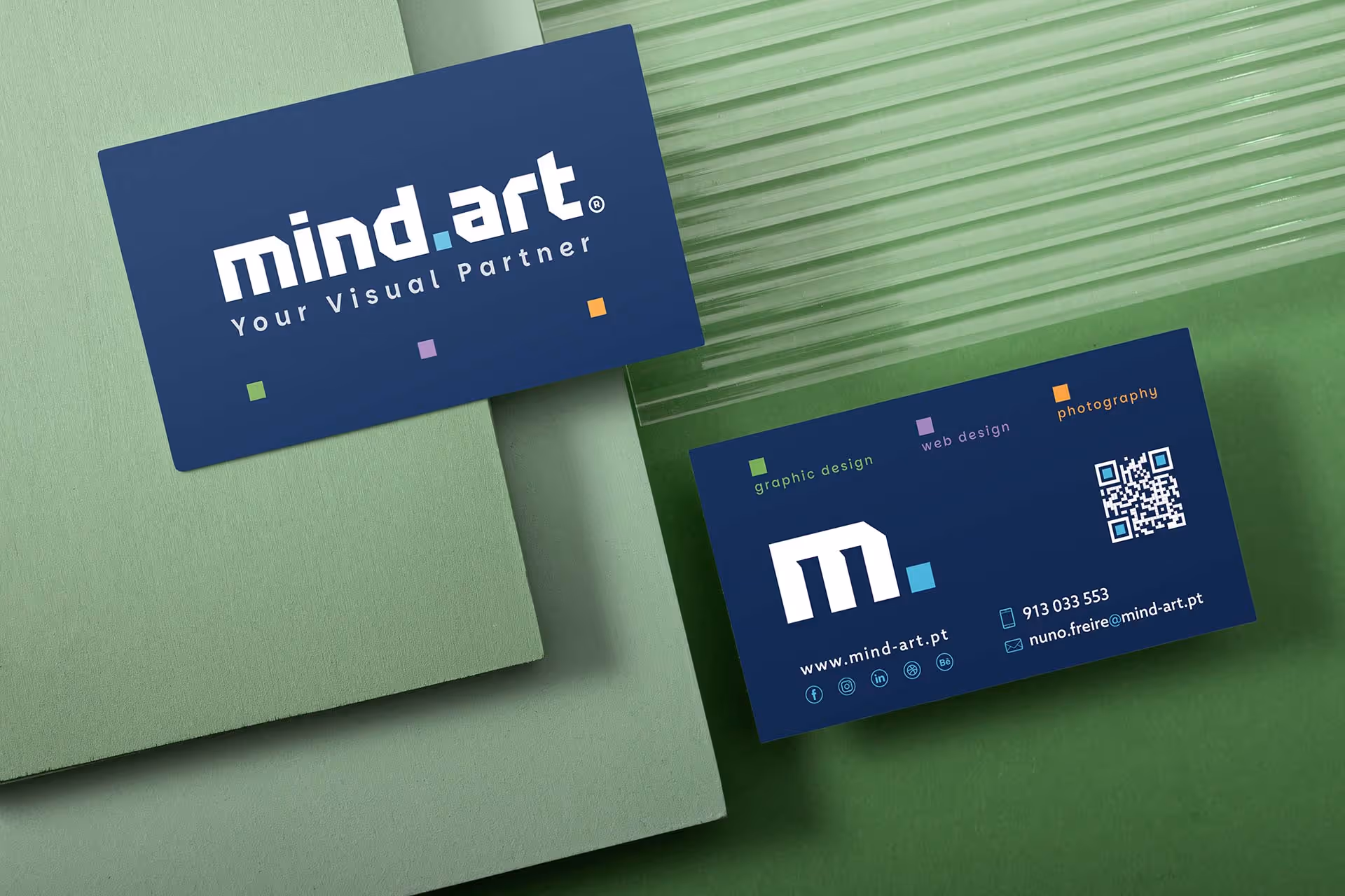 Dois cartões de visita azul escuro da empresa mind.art sobre fundos verdes, mostrando logotipo, serviços de design gráfico, web design e fotografia, e detalhes de contacto.