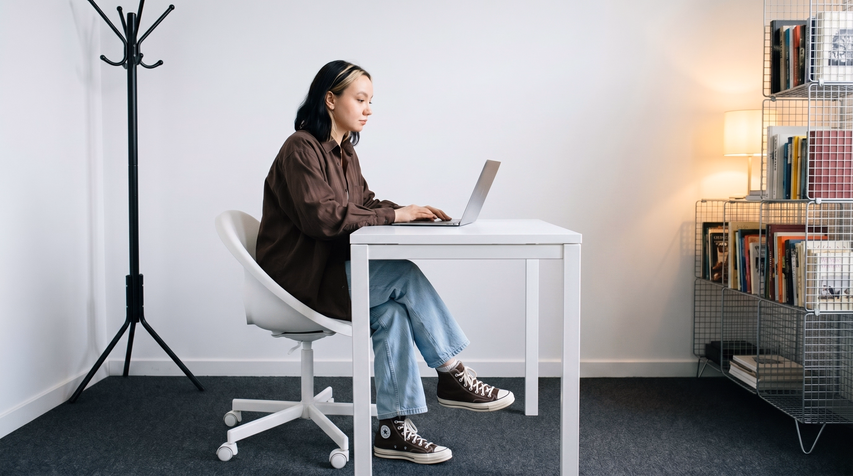 Person sitting at a white desk working on a laptop in a minimalist room with a bookshelf and lamp.
