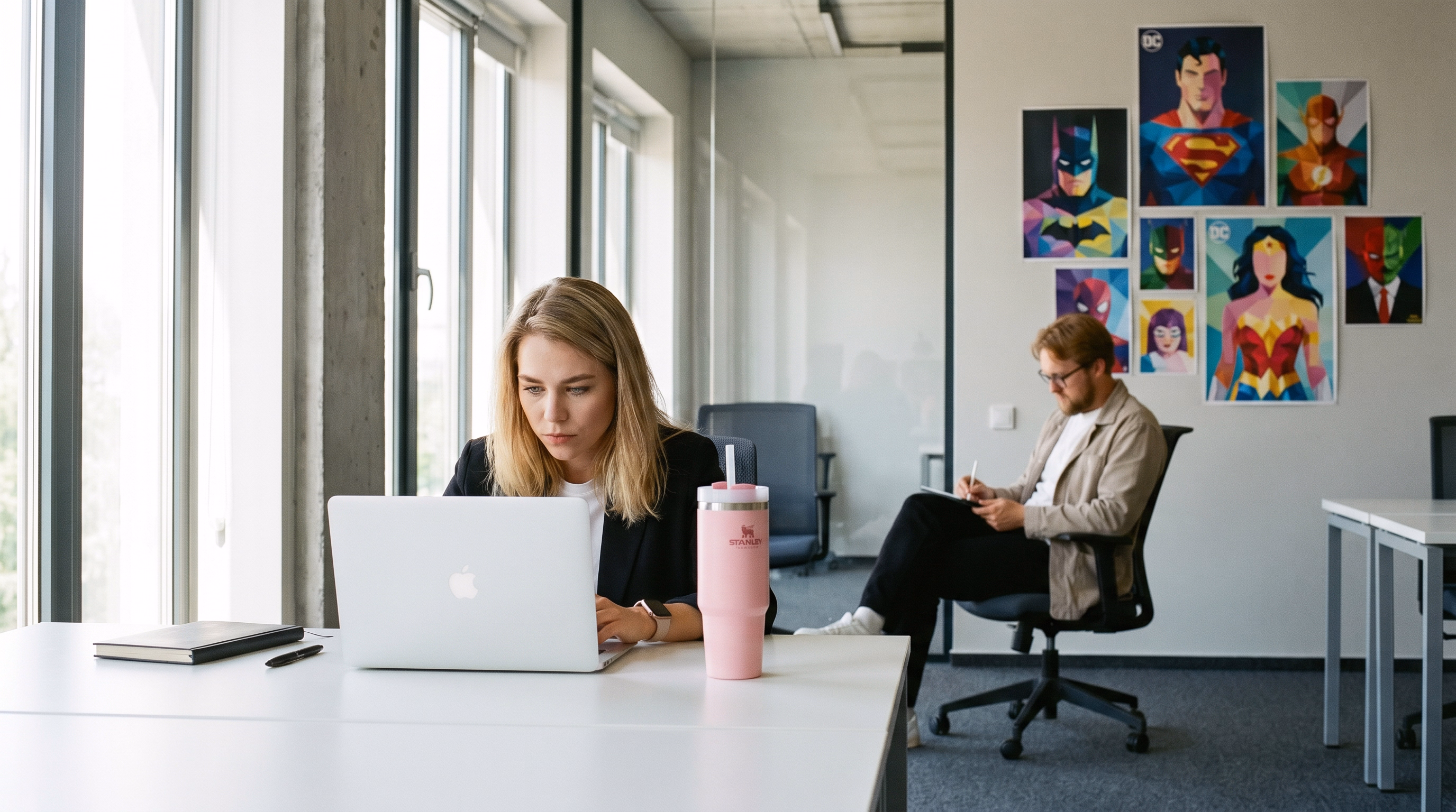 Woman focused on laptop at desk with pink Stanley tumbler; man in background writes on notepad in office.