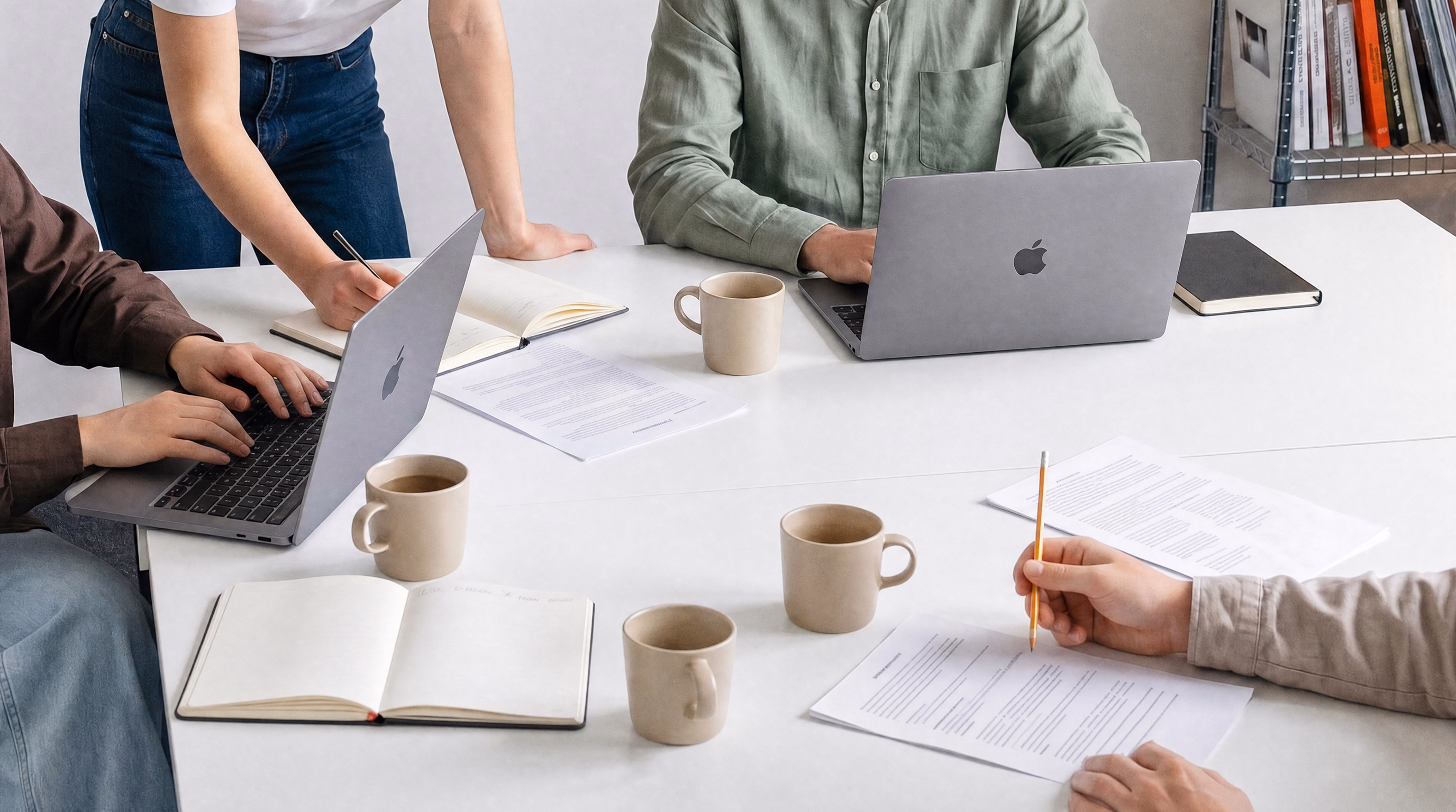People collaborating at a white table with laptops, notebooks, papers, and coffee mugs.