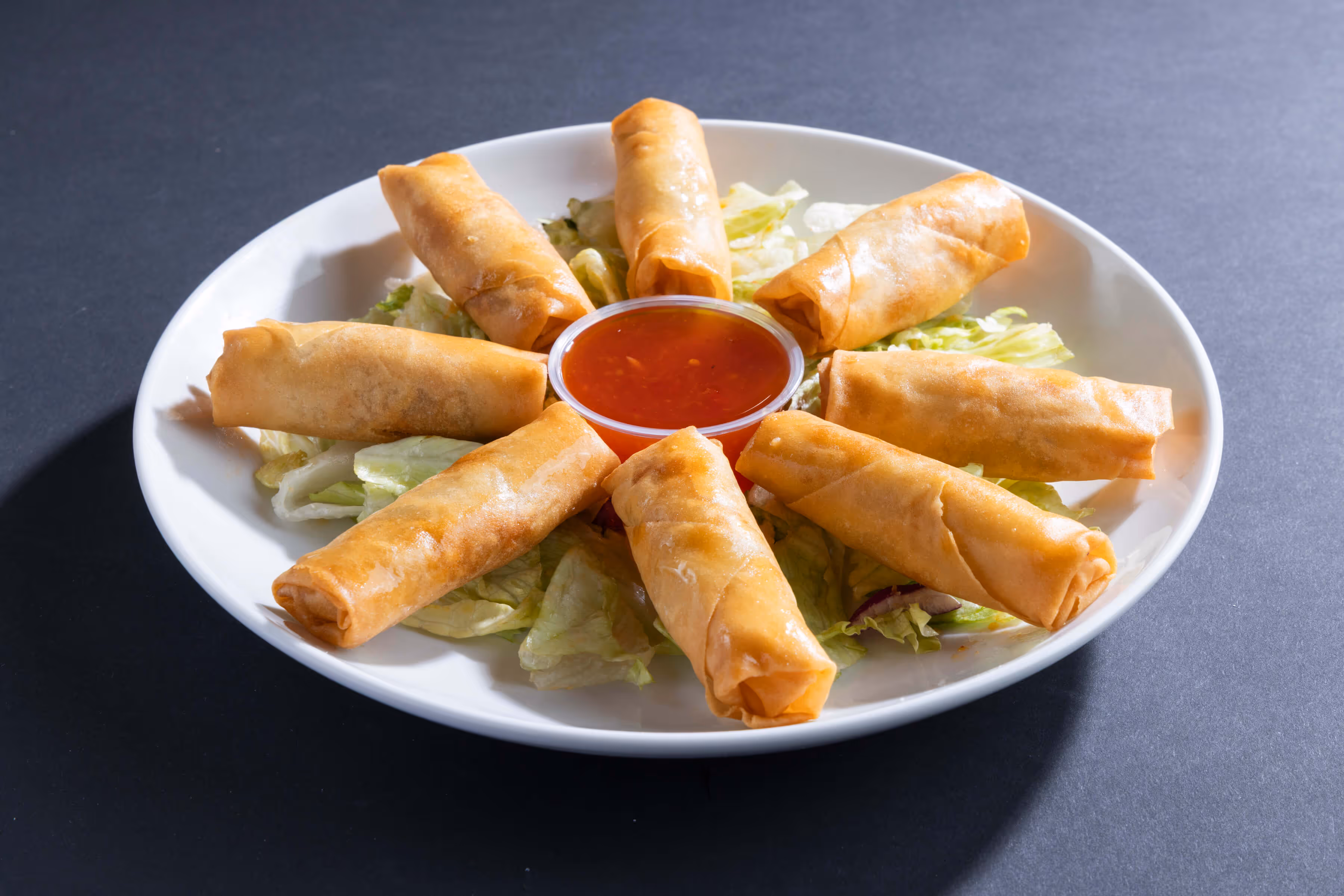 Plate of eight crispy vegetable spring rolls arranged around a bowl of red dipping sauce on lettuce.