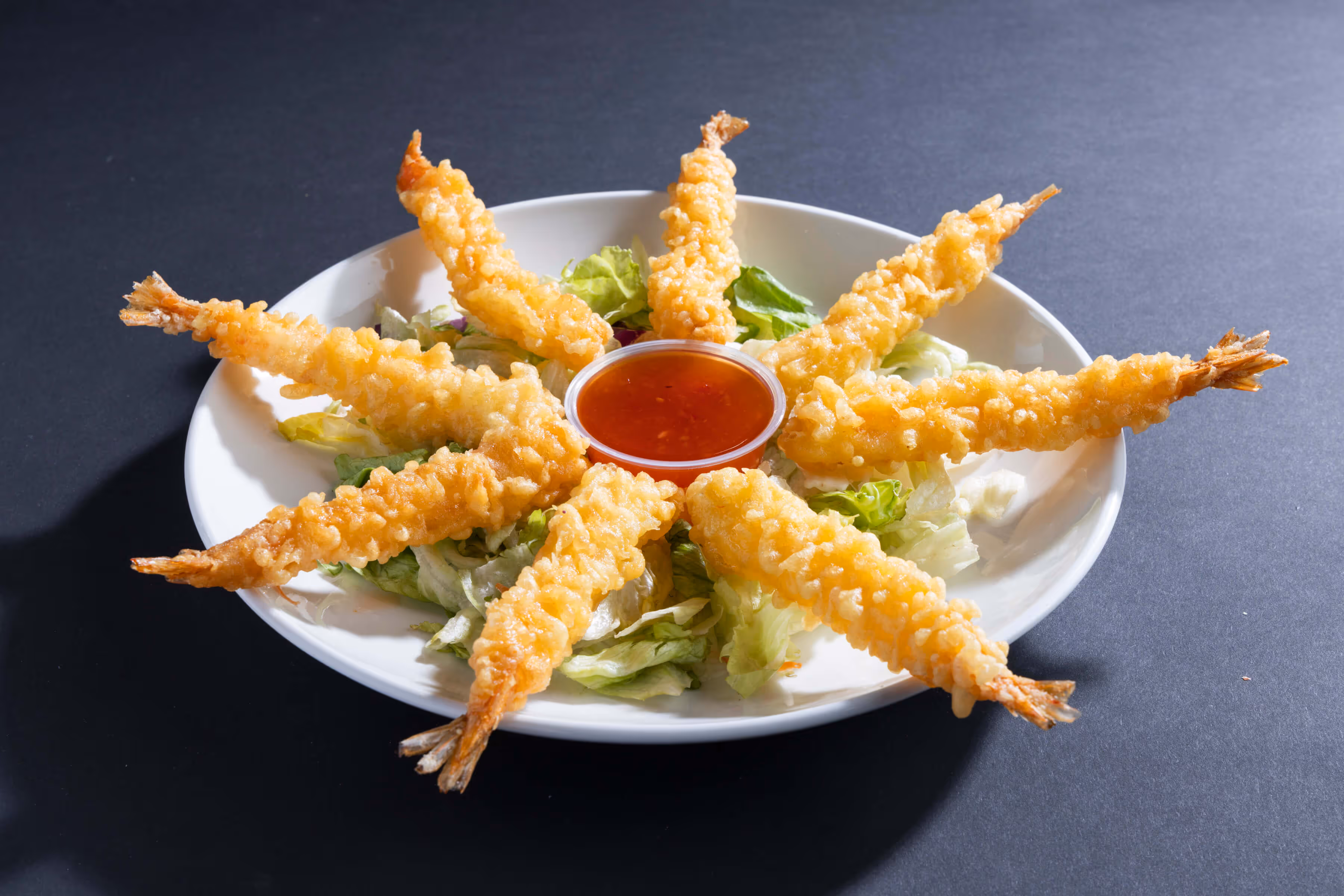 Plate of golden-brown shrimp tempura arranged in a circle on lettuce with a cup of red dipping sauce in the center.