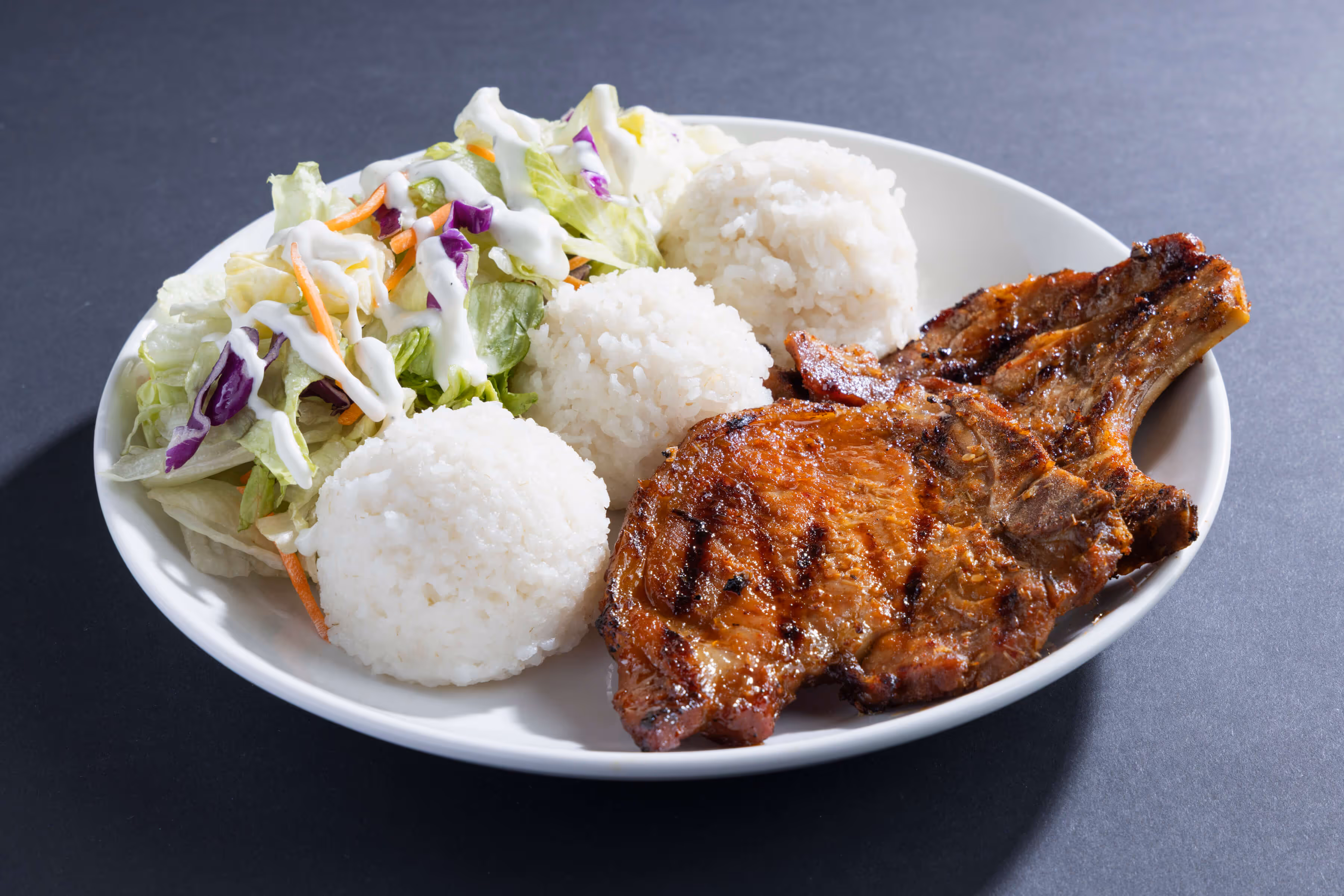 Grilled pork chop served with three scoops of white rice and a fresh salad topped with creamy dressing on a white plate.