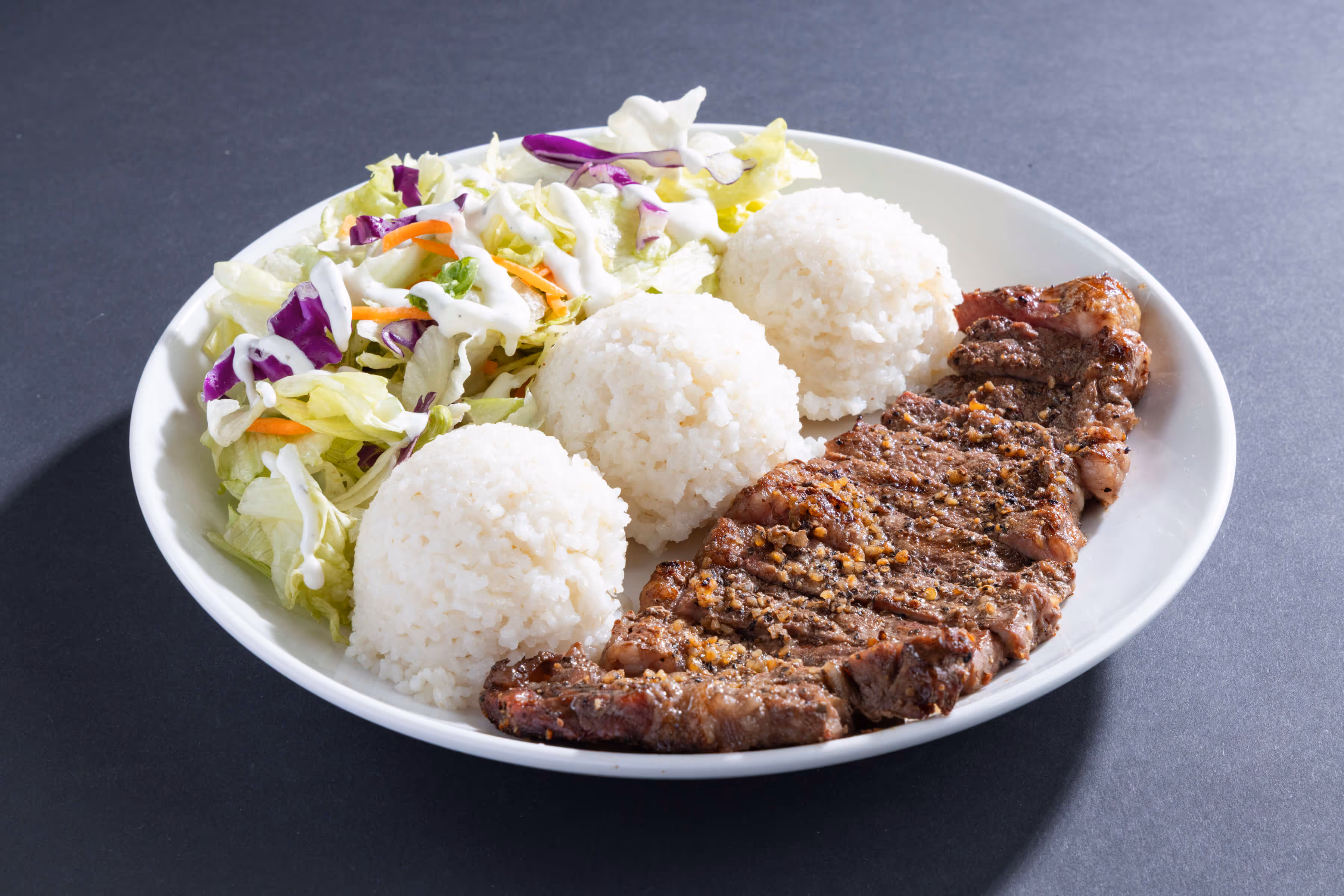 Plate with grilled steak, three scoops of white rice, and mixed salad topped with creamy dressing.