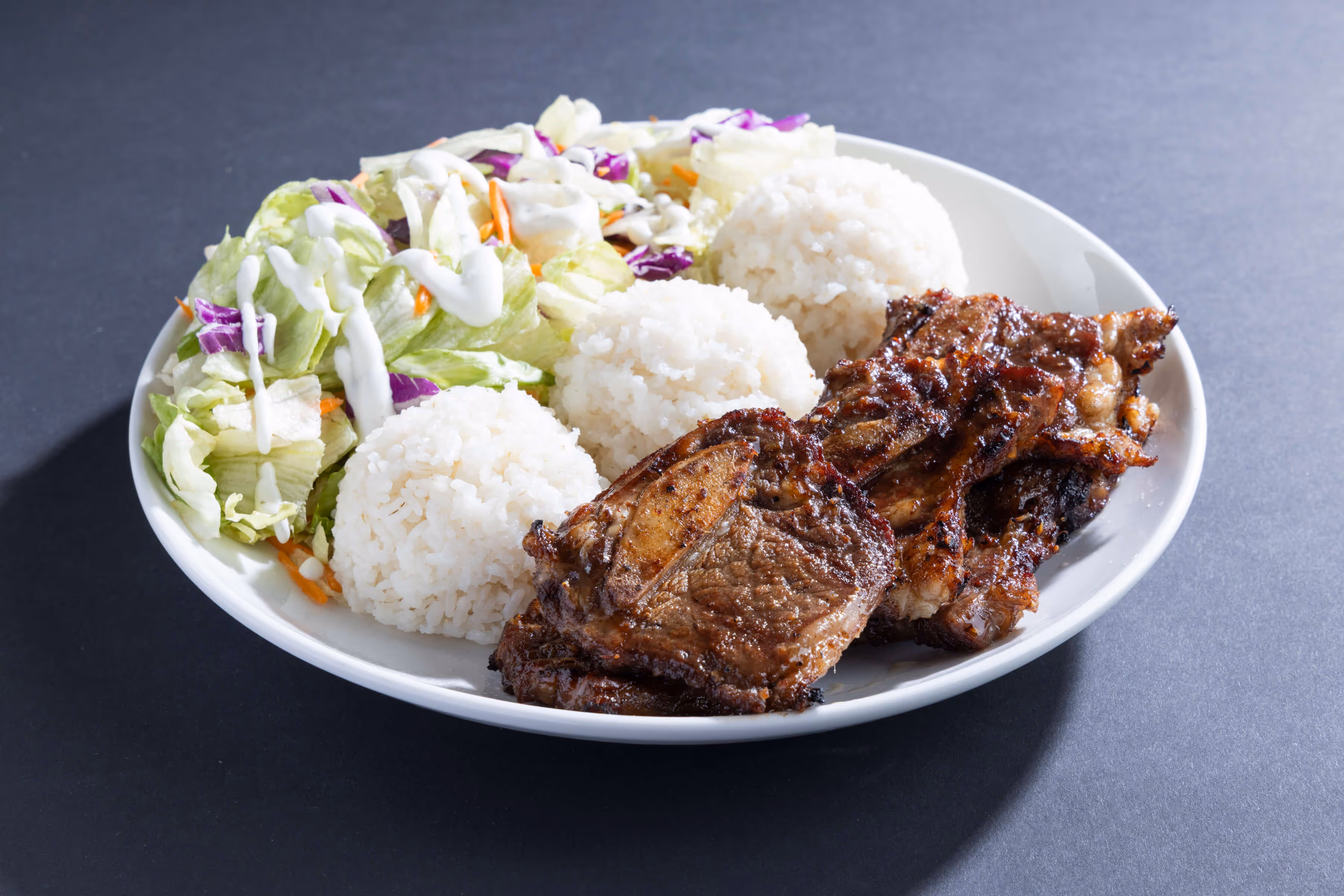Plate with grilled beef ribs, three scoops of white rice, and a side salad with lettuce, purple cabbage, and creamy dressing.