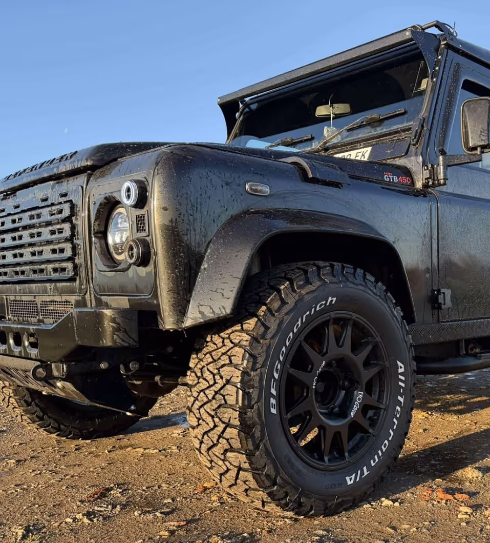 Close-up of a black off-road vehicle with BFGoodrich All-Terrain T/A tires parked on dirt under clear blue sky.