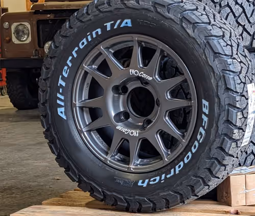 Close-up of a BFGoodrich All-Terrain T/A tire mounted on a gray EVO Corse alloy wheel, displayed on a wooden pallet in a garage.