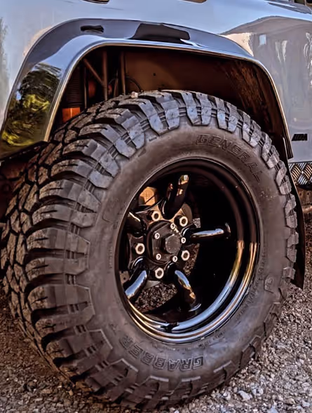 Close-up of a large off-road tire with aggressive tread mounted on a black rim, attached to a dark vehicle on gravel.