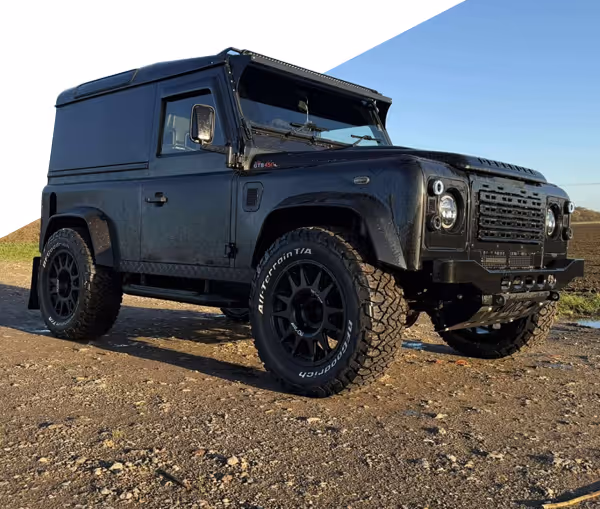 Black customized Land Rover Defender parked on rocky ground under clear blue sky with Devon Defenders logo above.