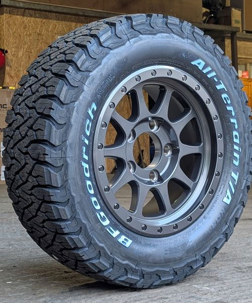 Close-up of a BFGoodrich All-Terrain T/A tire mounted on a gray EVO Corse alloy wheel, displayed on a wooden pallet in a garage.