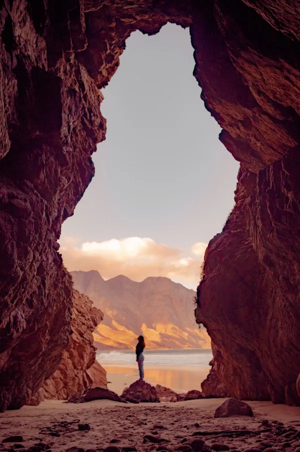 Personne debout sur un rocher à l'entrée d'une grotte regardant vers la mer et des montagnes au coucher du soleil.
