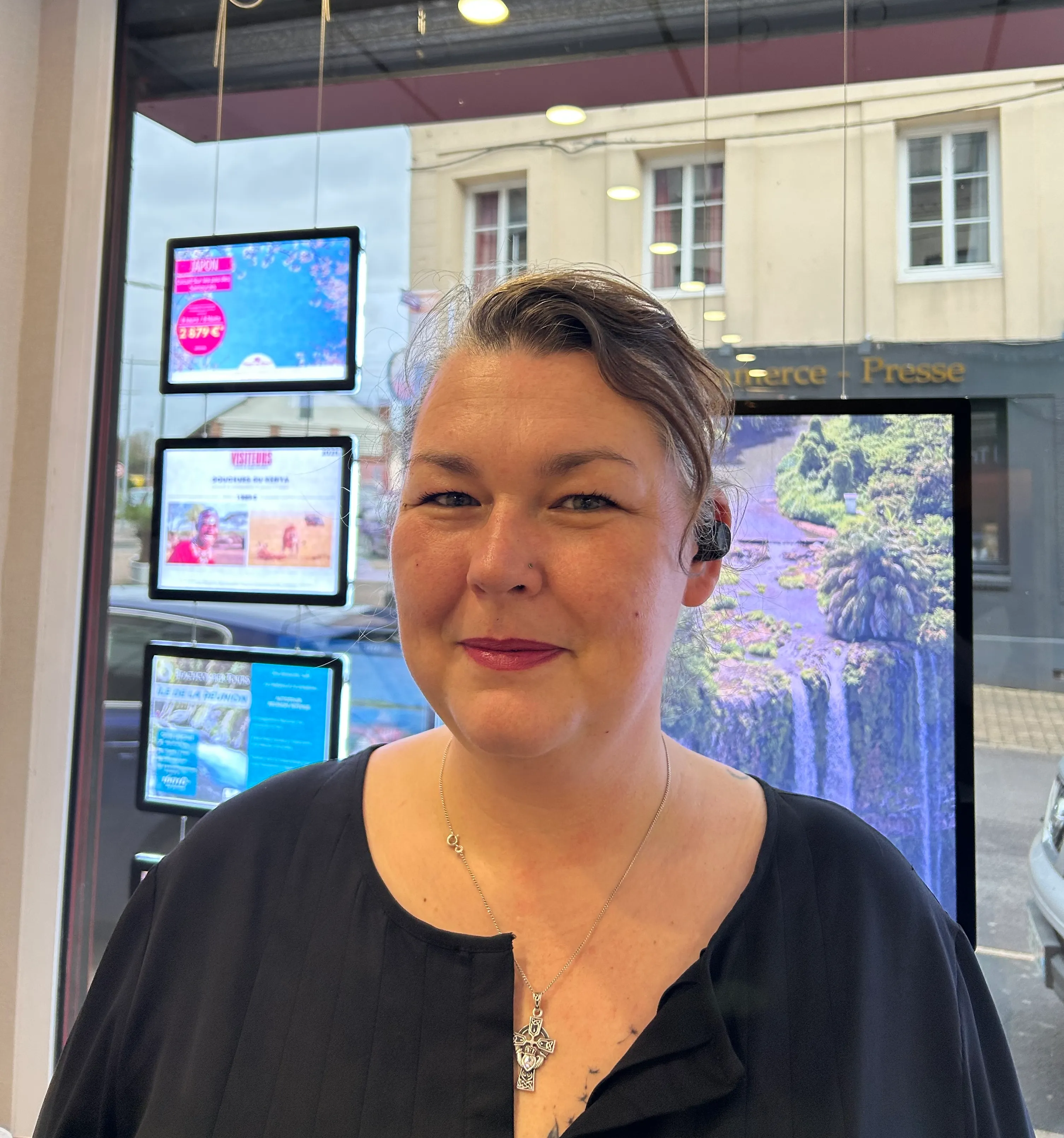 Portrait d'une femme souriante portant un haut noir et un collier croix, avec une oreillette noire, devant une vitrine de magasin avec affichages lumineux.