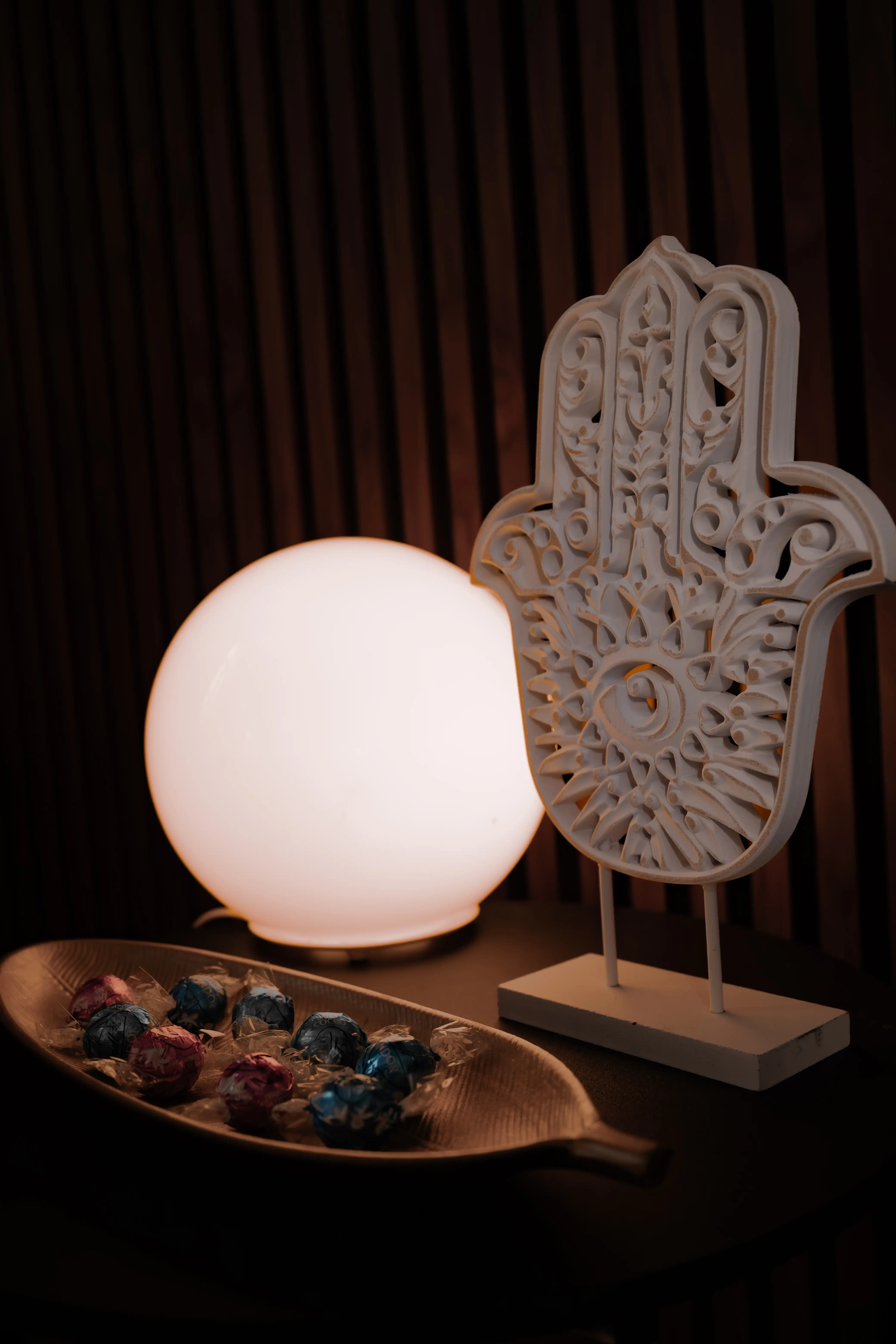 Eine kunstvoll verzierte weiße Hamsa-Hand, beleuchtet von einer runden gelben Lampe, daneben eine Schale mit bunten Bonbons vor einem dunkel gestreiften Hintergrund.