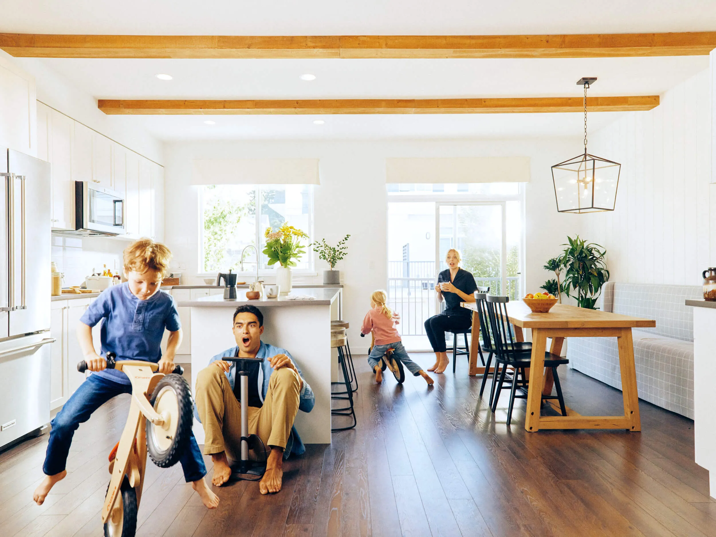 A family enjoying indoors with two children playing on balance bikes and a woman seated at the dining table drinking a beverage in a bright kitchen.