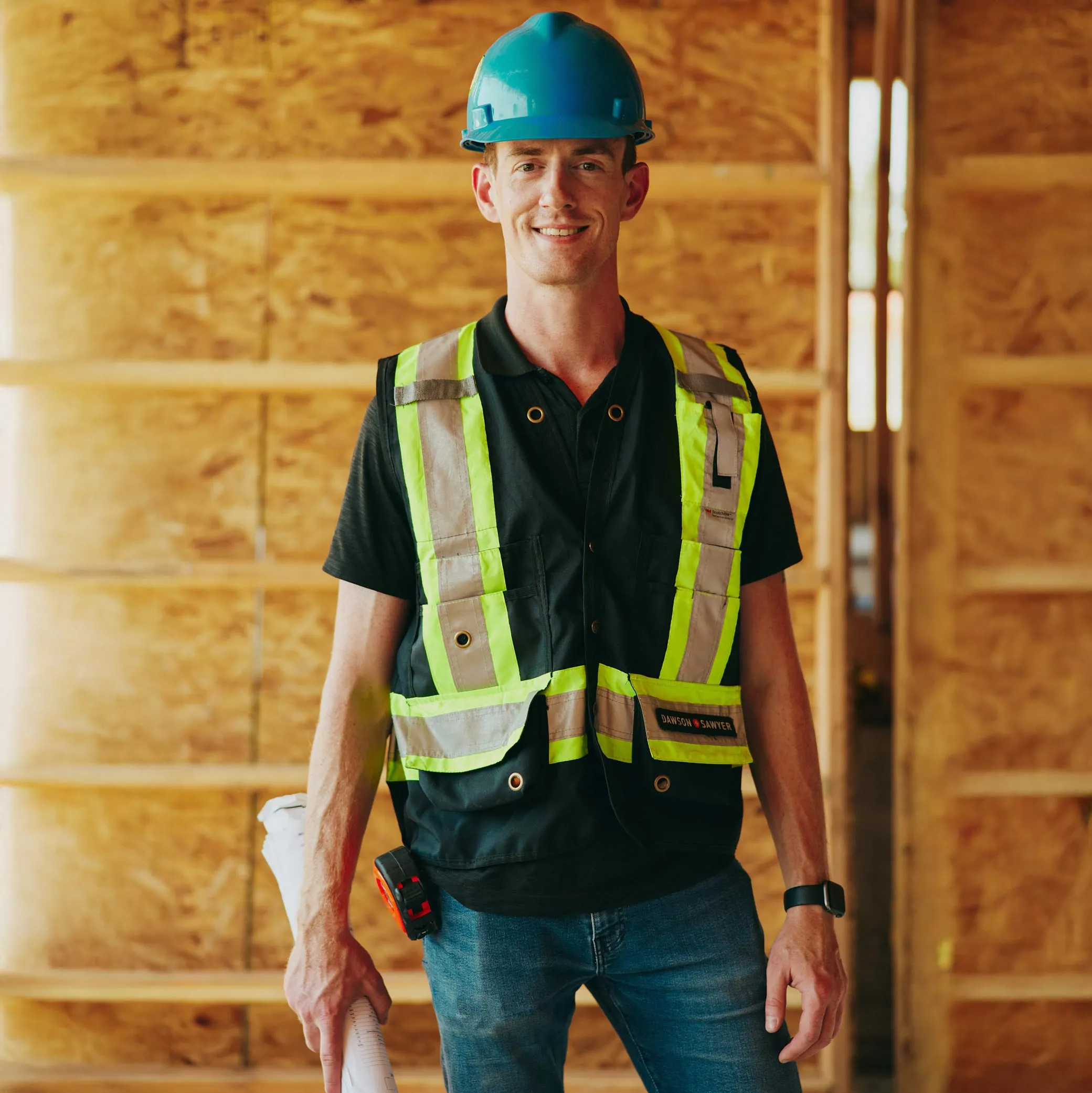Smiling construction worker wearing a blue hard hat and reflective safety vest holding rolled-up blueprints.