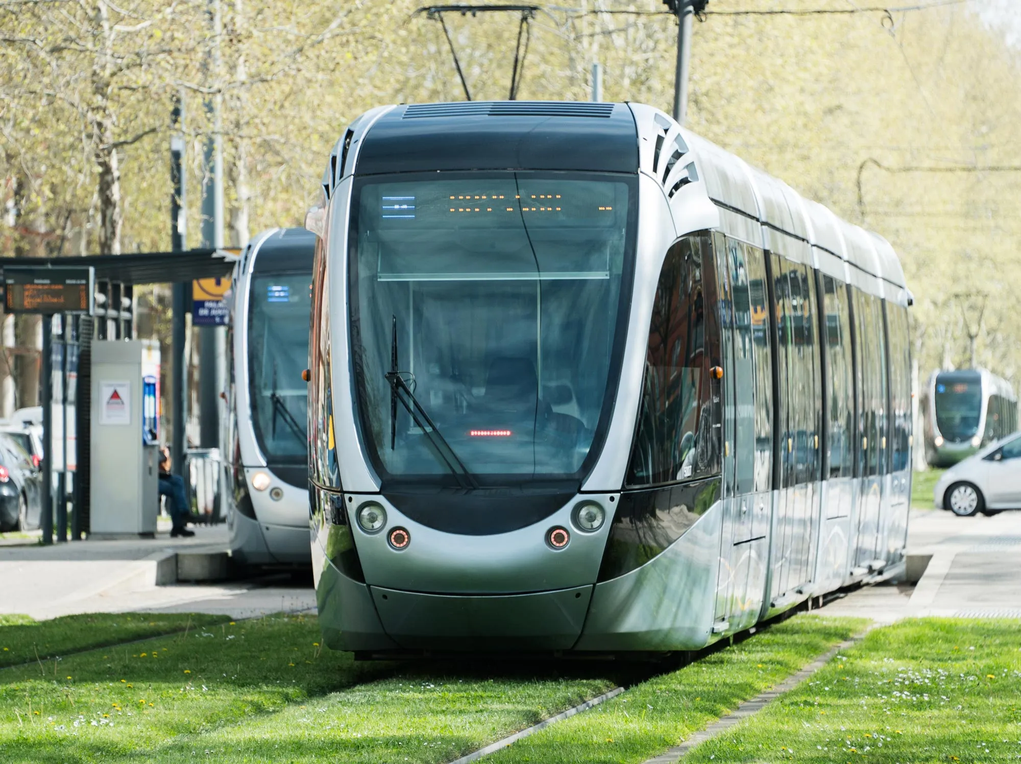 Modern silver tram moving on grass-covered tracks with another tram and passengers at a station in the background.
