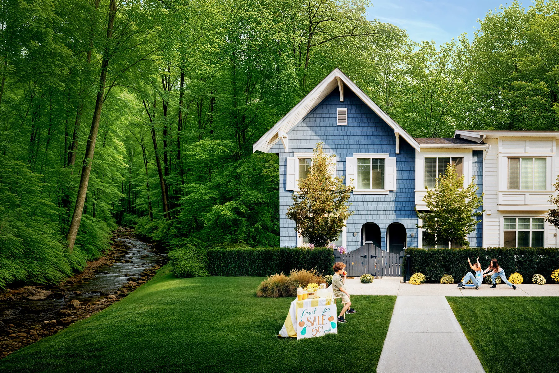 Two children playing on a skateboard near a blue and white house by a forest with a stream, while another child sells fruit at a stand in the front yard in bear creek surrey townhomes