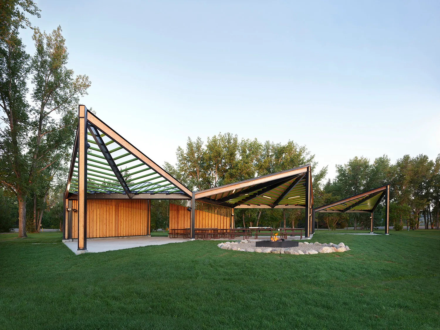 Modern wooden and metal pavilion with triangular roof sections in a grassy park with a fire pit in front at Queens in Surrey