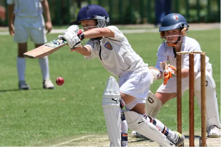Young cricket player in white uniform and helmet batting, with wicketkeeper and stumps behind on green field at Queens condos in Surrey