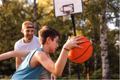 Young boy holding a basketball with a man watching him in an outdoor basketball court surrounded by trees at Queens
