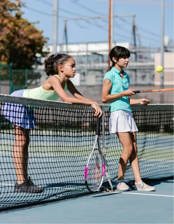 Two girls leaning on a tennis net on an outdoor court, one holding a tennis racquet and watching a tennis ball in mid-air at Queens in surrey