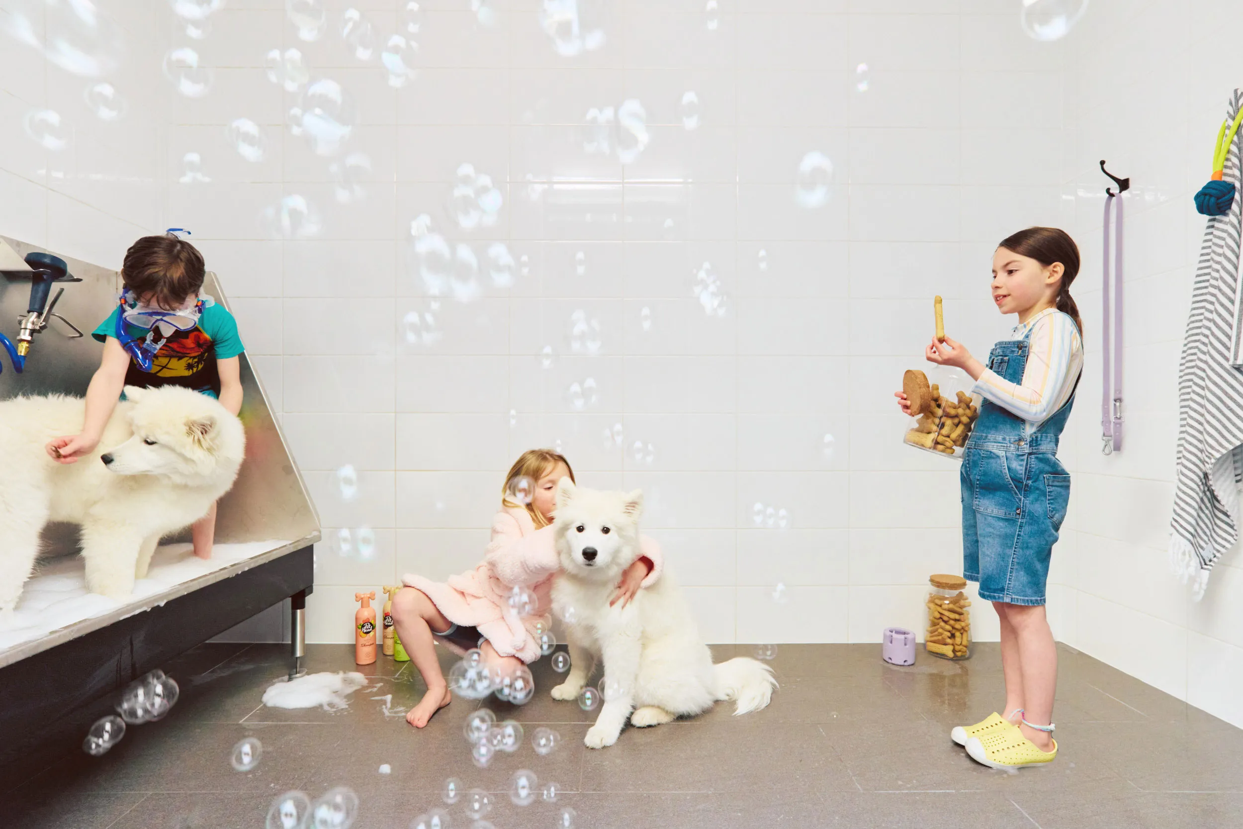 Three children interact with two fluffy white dogs in a clean, tiled room filled with bubbles, one child holds a jar of dog treats at Fleetwood Village Condos in Surrey