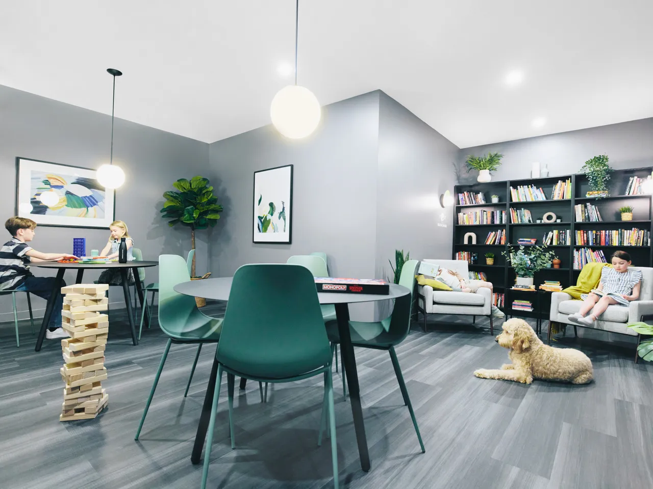 Children sitting at tables playing board games and reading in a modern, gray-walled room with bookshelves and a dog lying on the floor at Fleetwood Village in Surrey