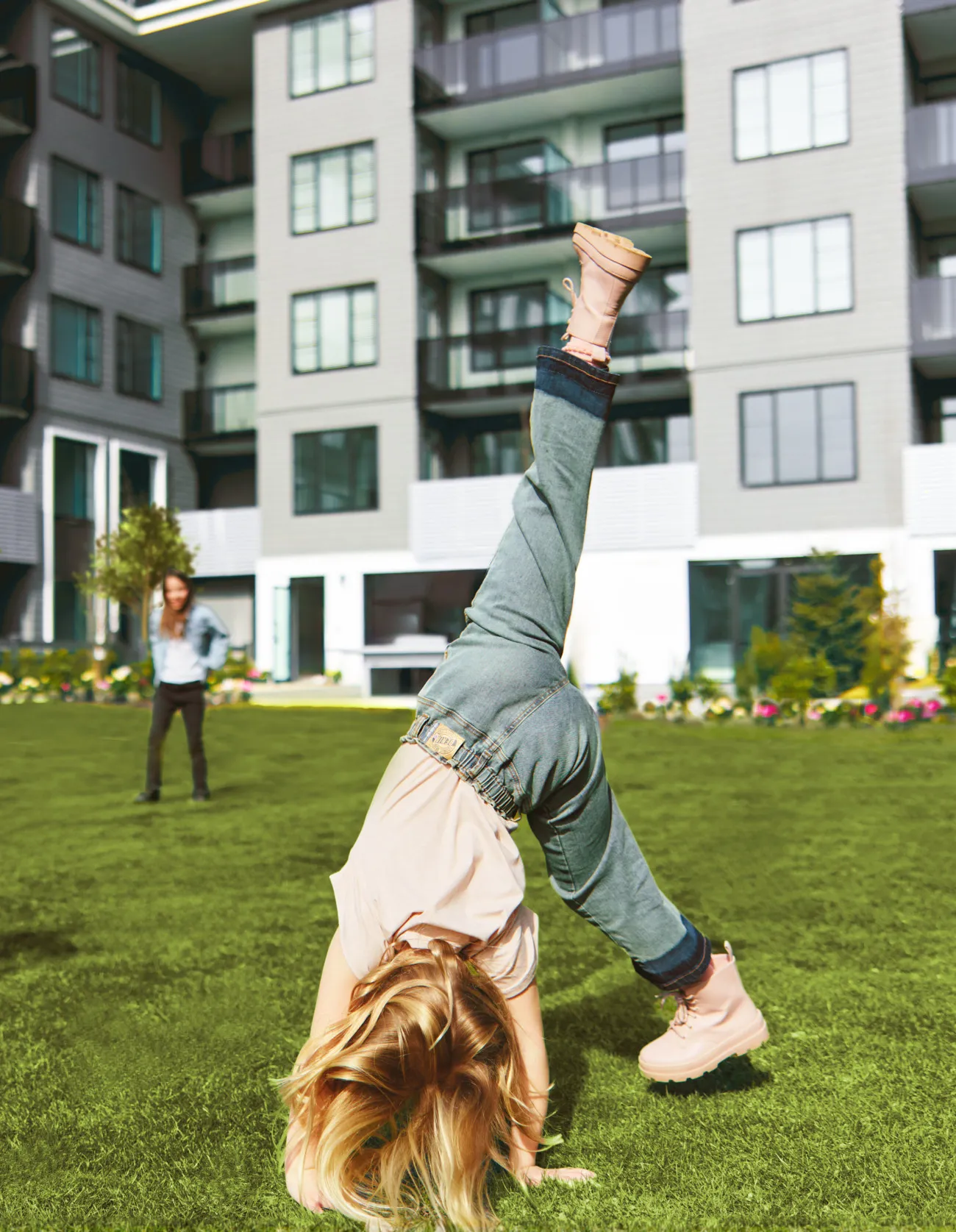 Child performing a handstand on grass in front of modern apartment buildings with another child standing in the background at Fleetwood Village 2 Park