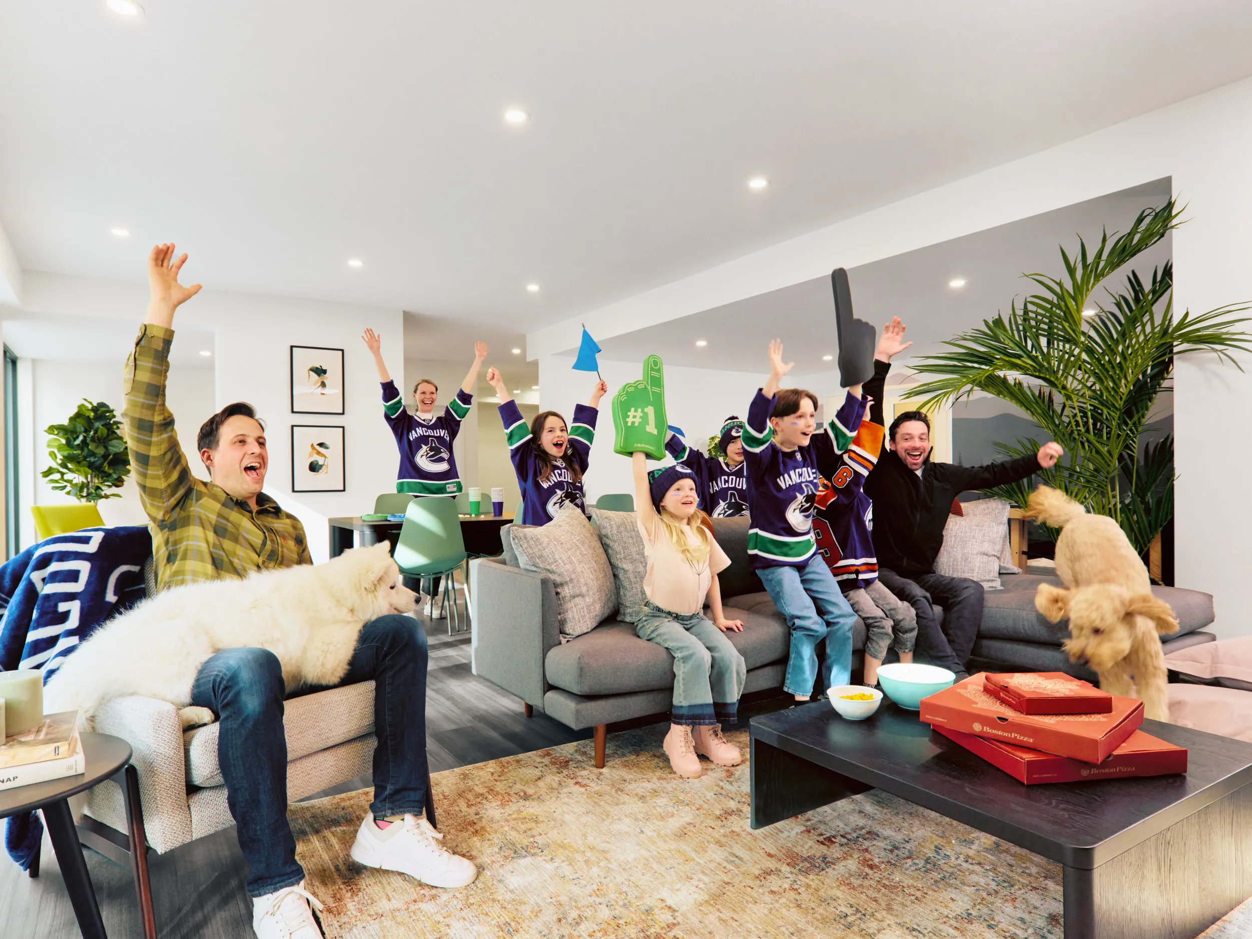 Group of people and children in Vancouver Canucks jerseys cheering enthusiastically in a living room with two dogs and pizza boxes on the coffee table at the amenity lounge for Fleetwood Village 2