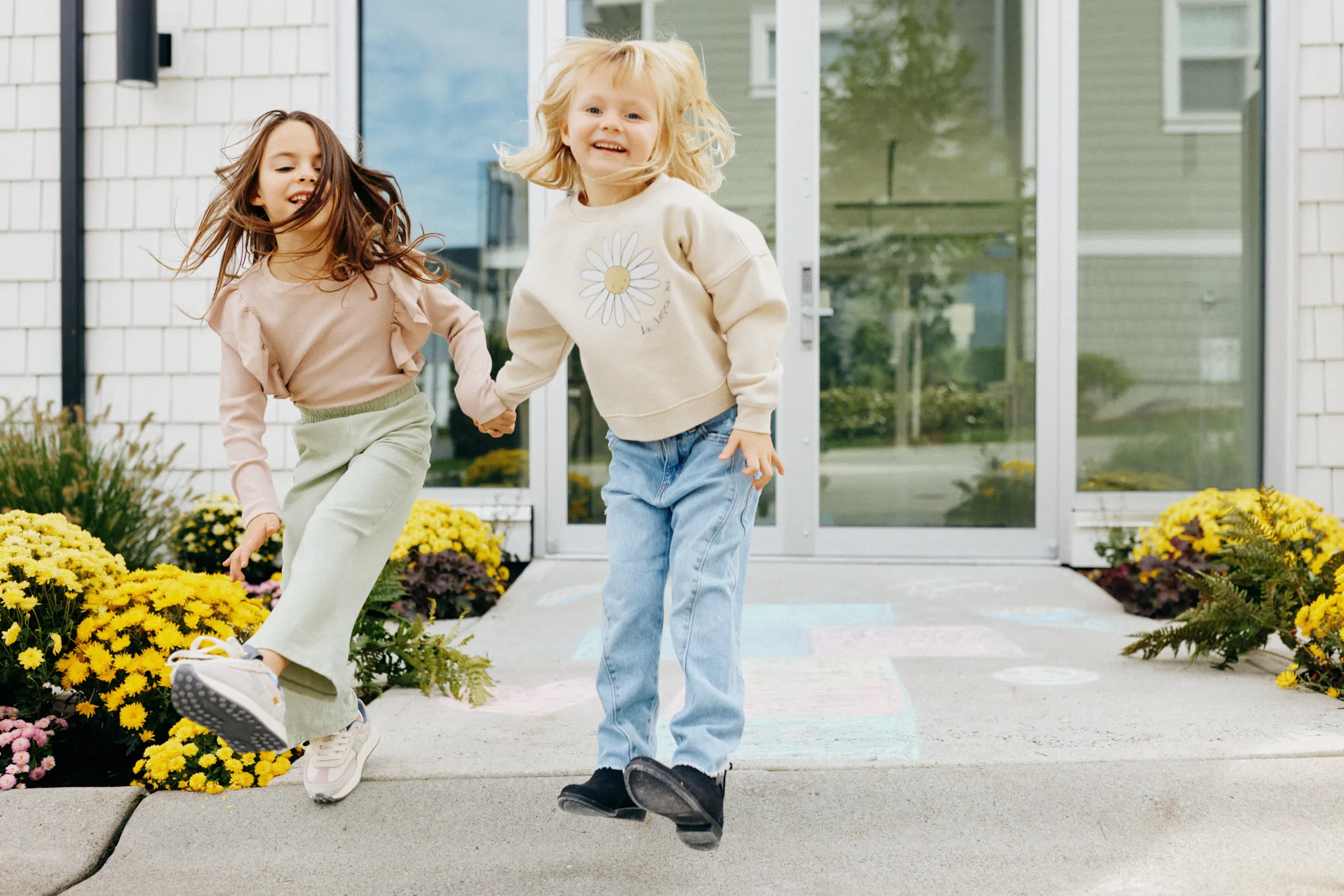 Two young girls holding hands and joyfully jumping on a sidewalk in front of a house with yellow flowers at amenity building for dawson + sawyer