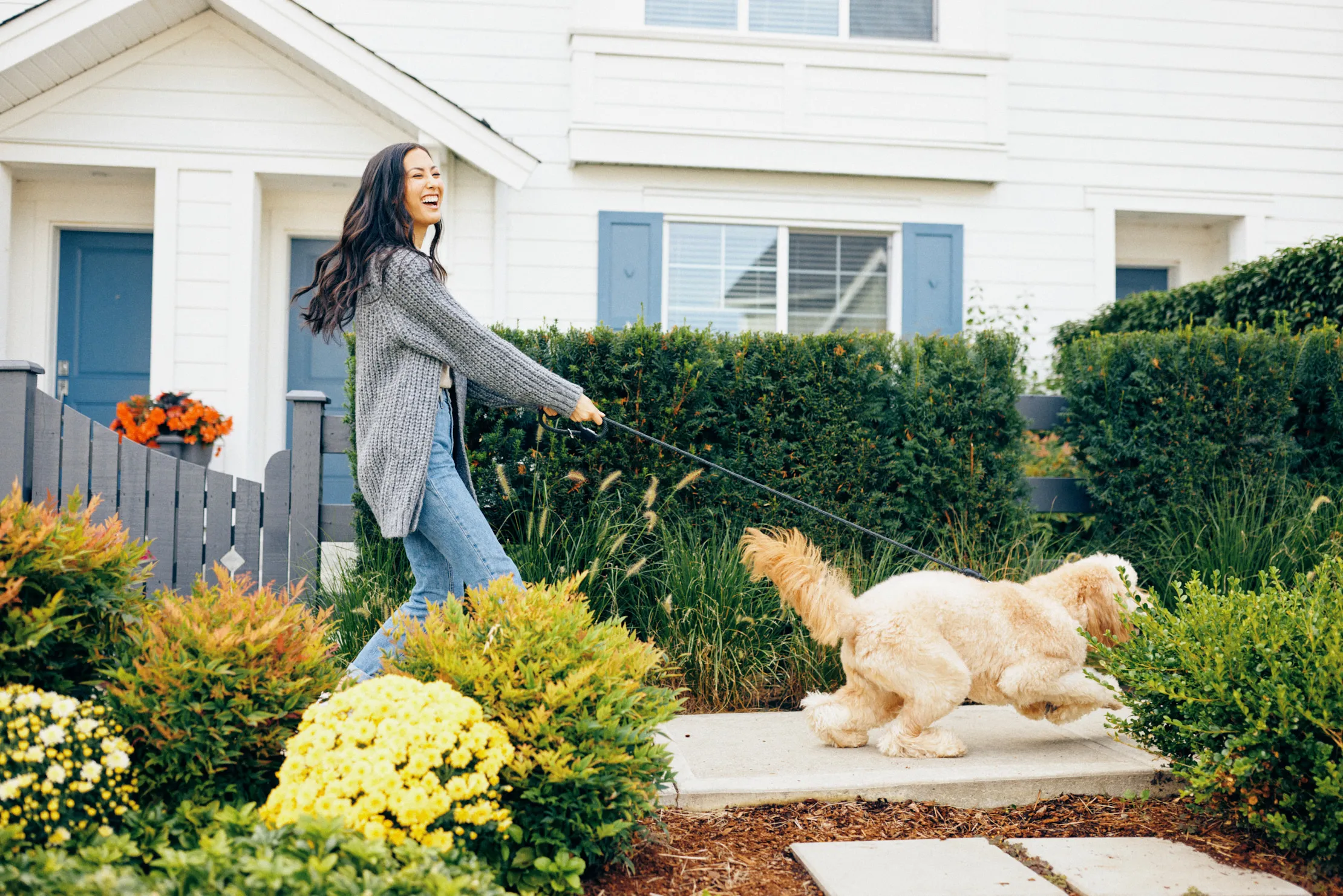 Smiling woman in a gray sweater walking a fluffy light-colored dog on a leash in a garden in front of a white house at Fleetwood Village townhomes