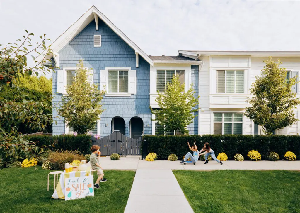 Two children playing on the sidewalk in front of a blue and white house with a lemonade stand on the grass nearby townhomes by dawson + sawyer