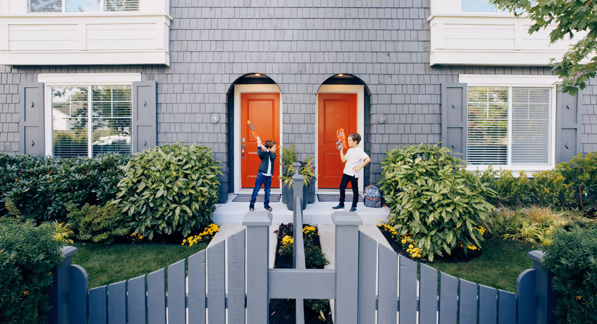 Two boys playing with toys facing each other on the front steps of adjacent houses with red doors and gray siding at Fleetwood Village Townhomes