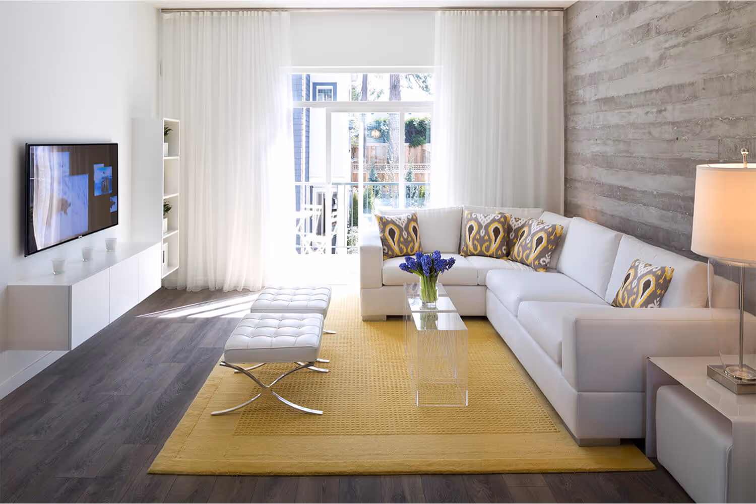 Modern living room with white sectional sofa, patterned yellow cushions, yellow rug, clear glass coffee table with purple flowers, white ottoman at Kings Landing Townhomes
