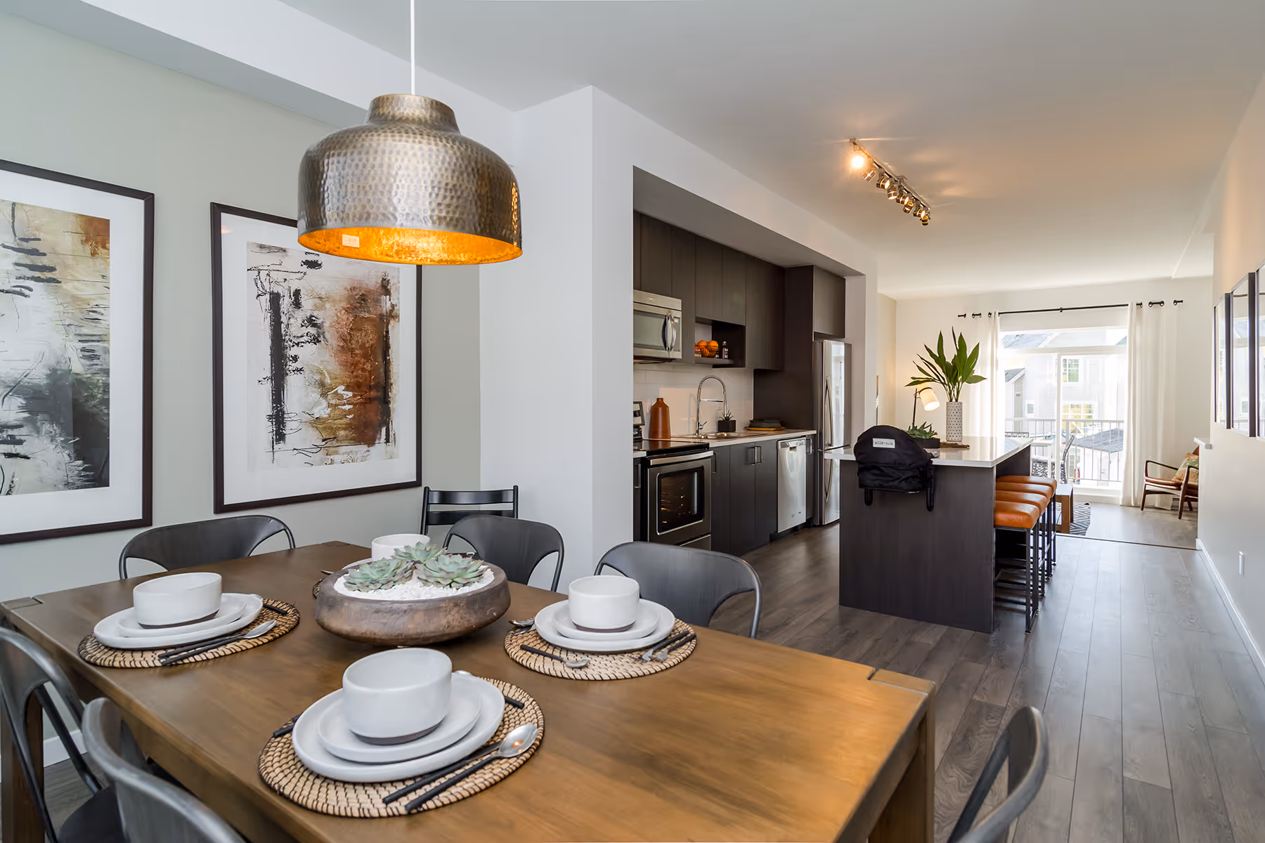 Modern dining area with wooden table set for four, pendant light, abstract wall art, and open kitchen with island and seating Guildford The Great