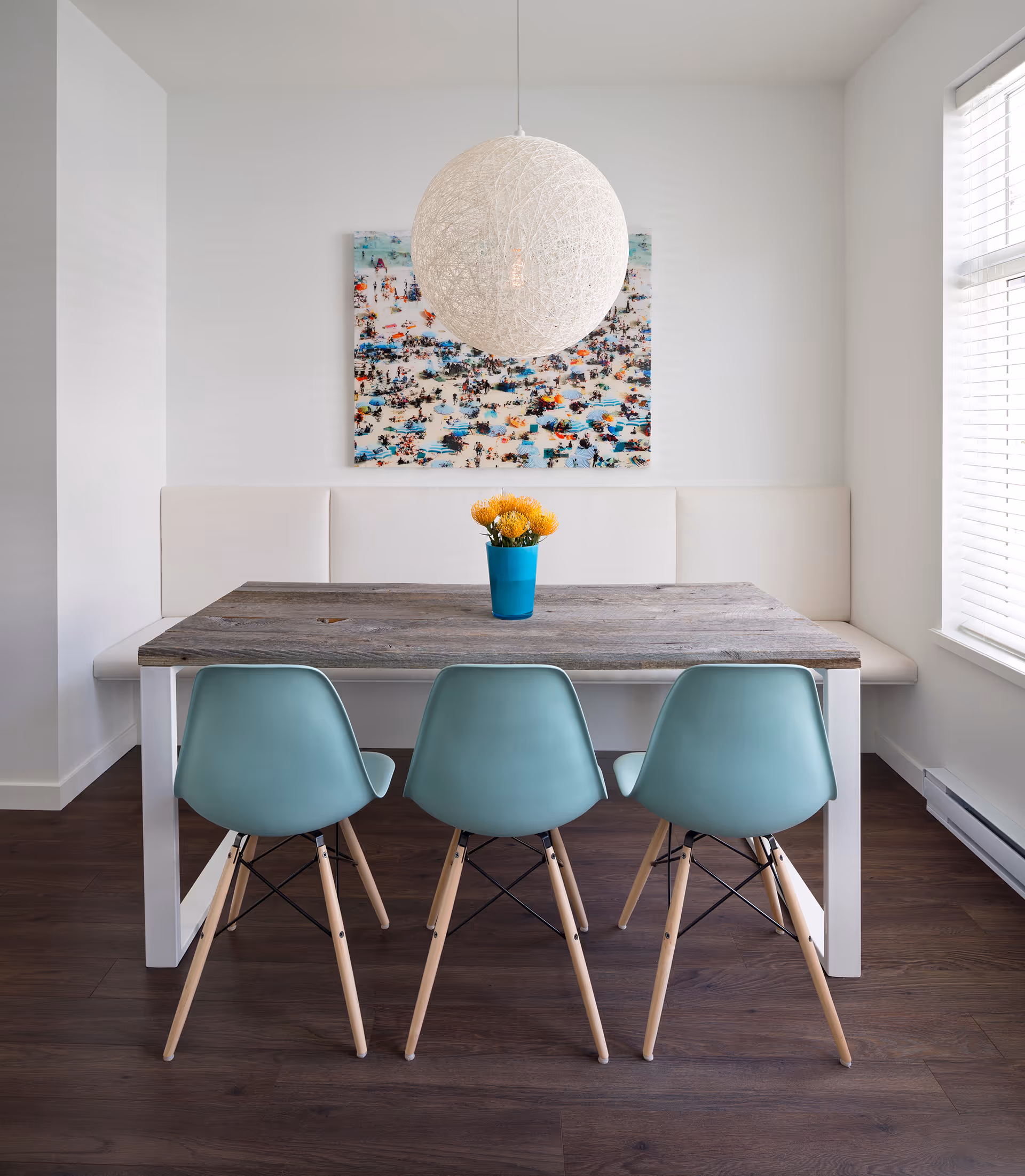 Modern dining area with a rectangular wooden table, three teal chairs with wooden legs, a white cushioned bench, a spherical white pendant light, and a colorful abstract painting on the wall.